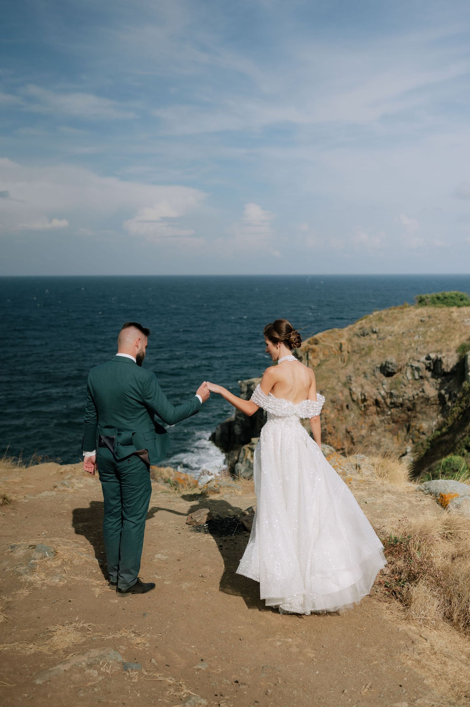 A playful wedding photo sequence captured on a rugged oceanside cliff under blue skies.