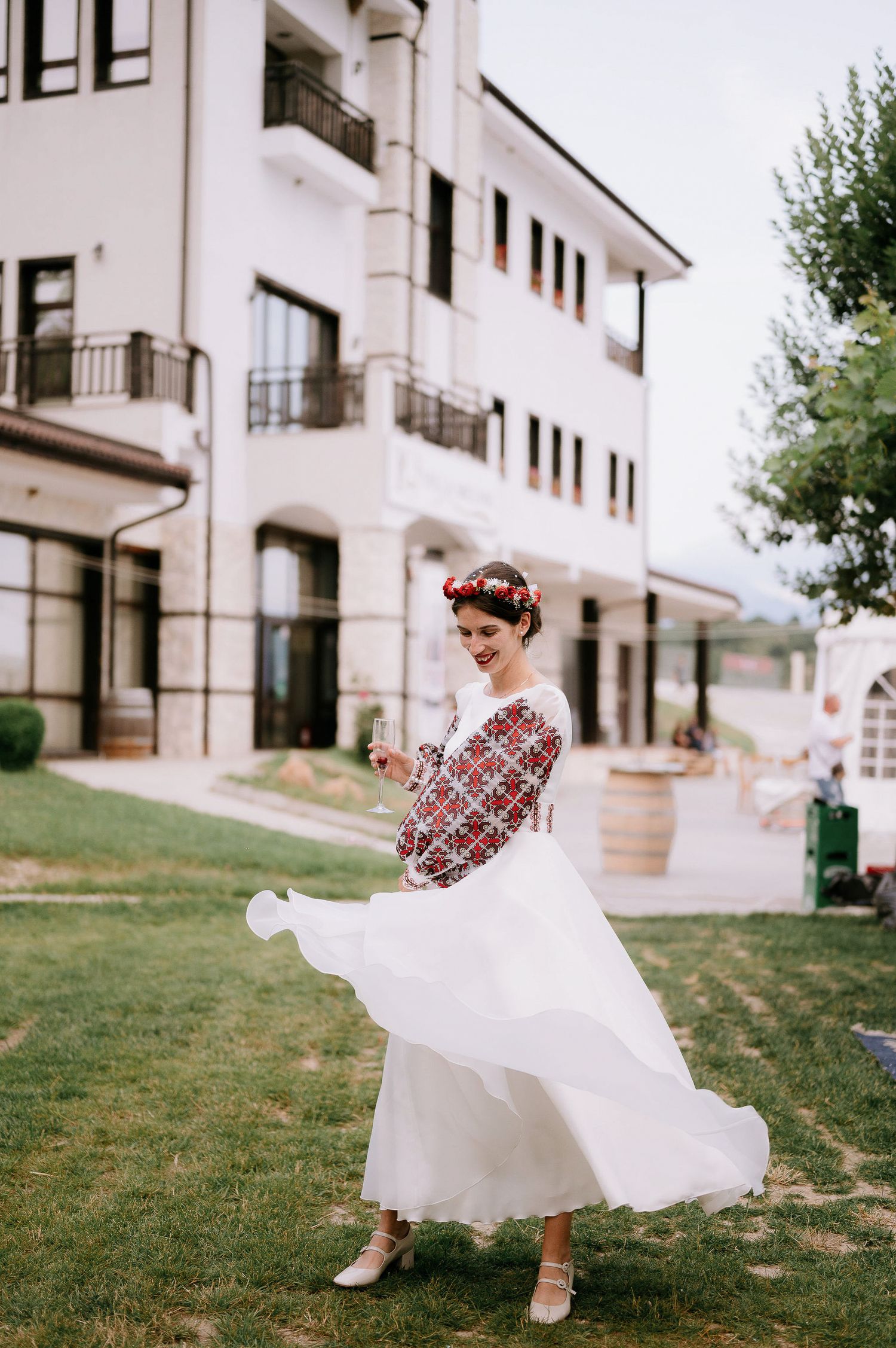 A white wedding dress with red embroidered sleeves twirls gracefully on a grassy lawn near a white building.