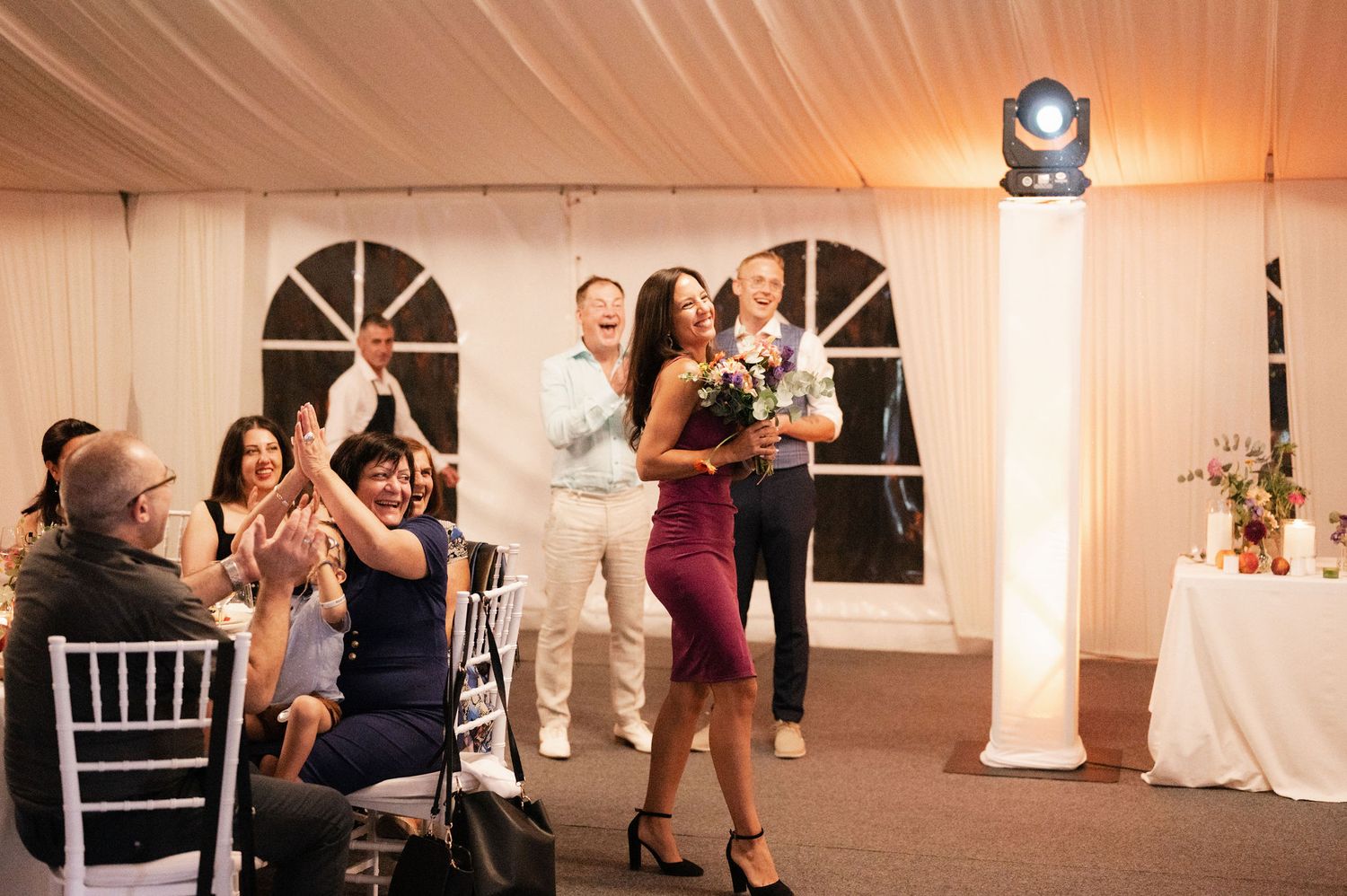 Wedding guests dance together on a wooden dance floor in a warmly lit reception tent with white walls and decor.