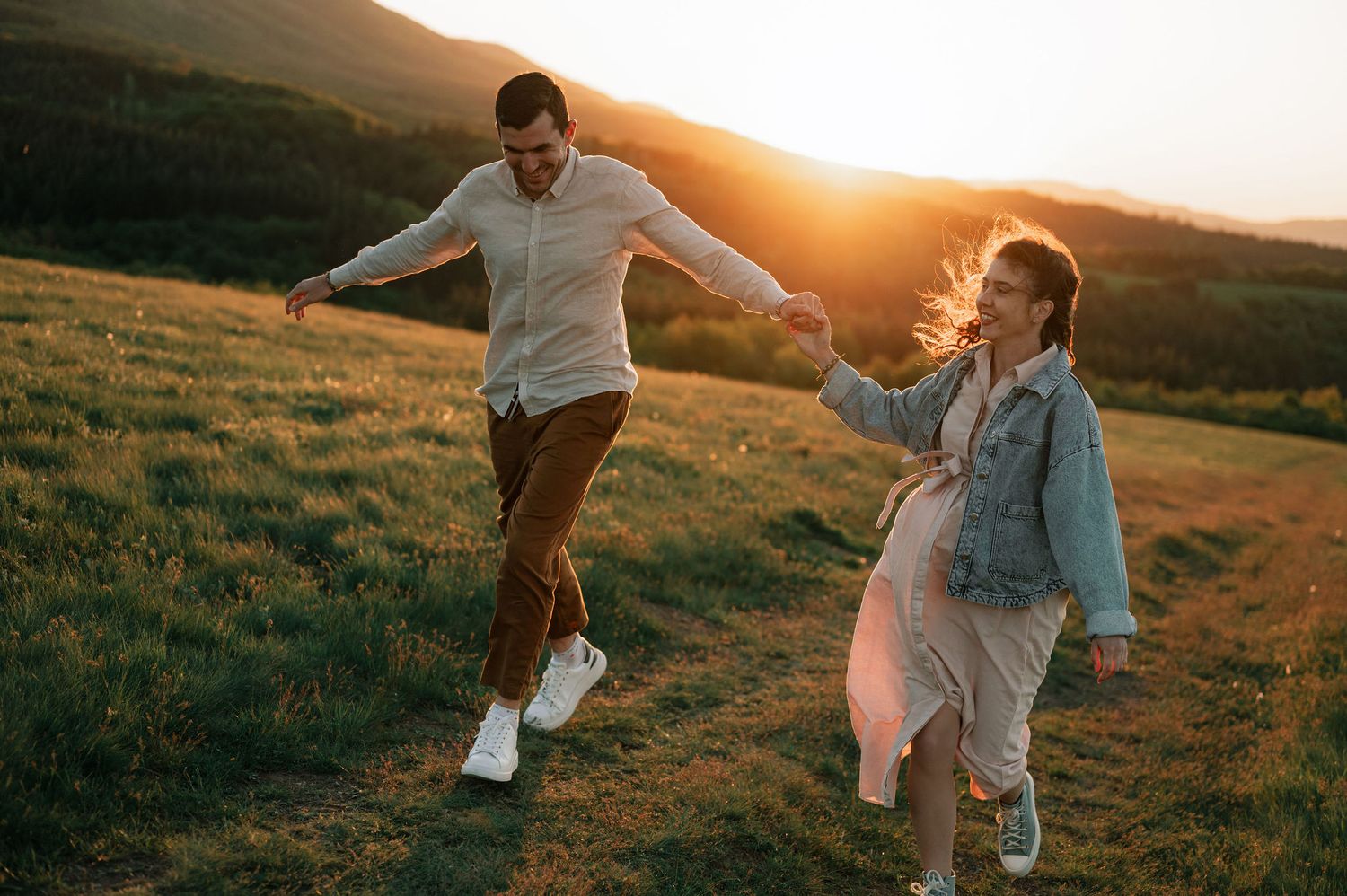 A couple playfully dances together in a grassy field during golden hour sunset.