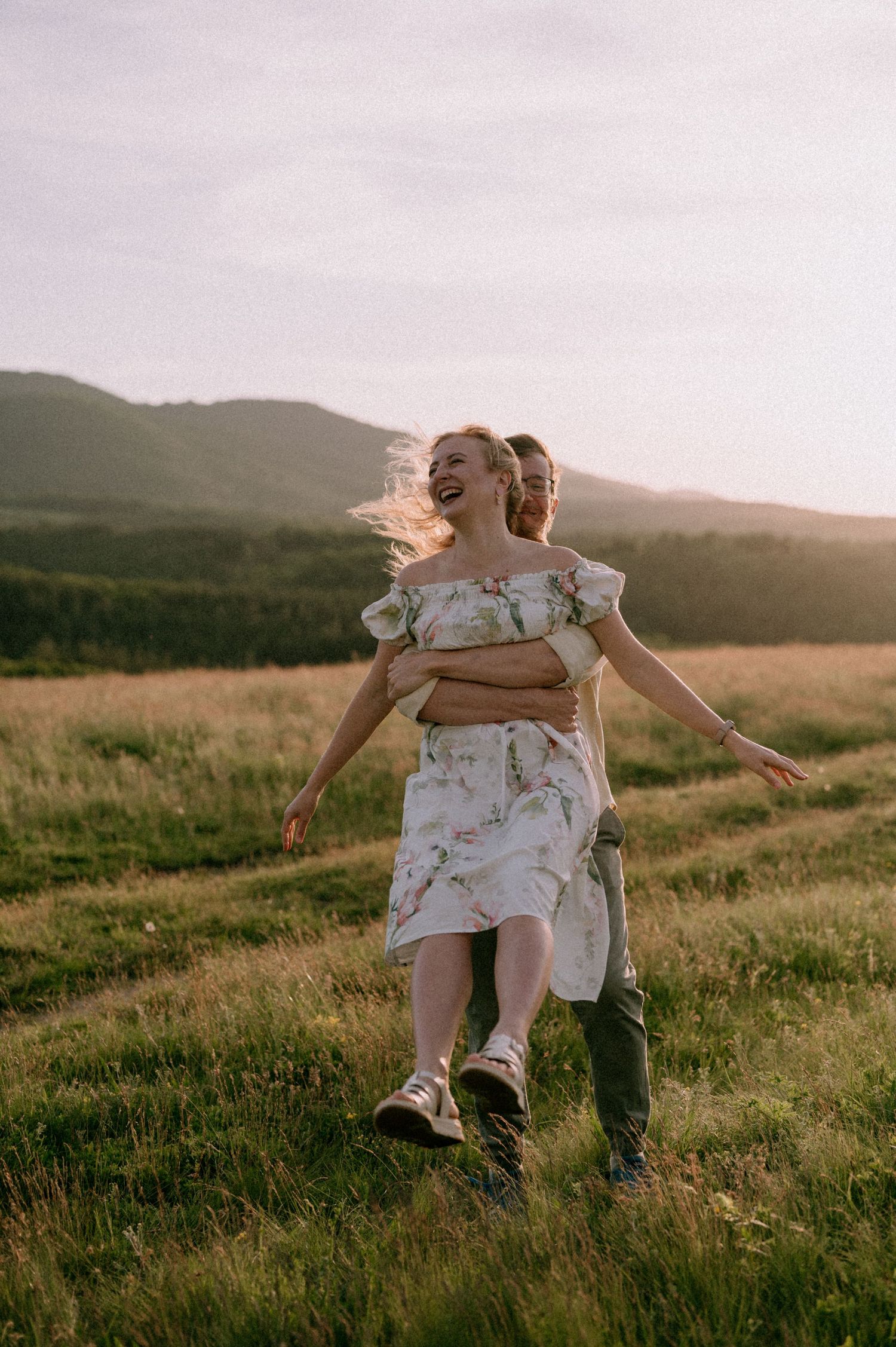 Playful couple enjoying piggyback rides in a scenic mountain field at dusk.