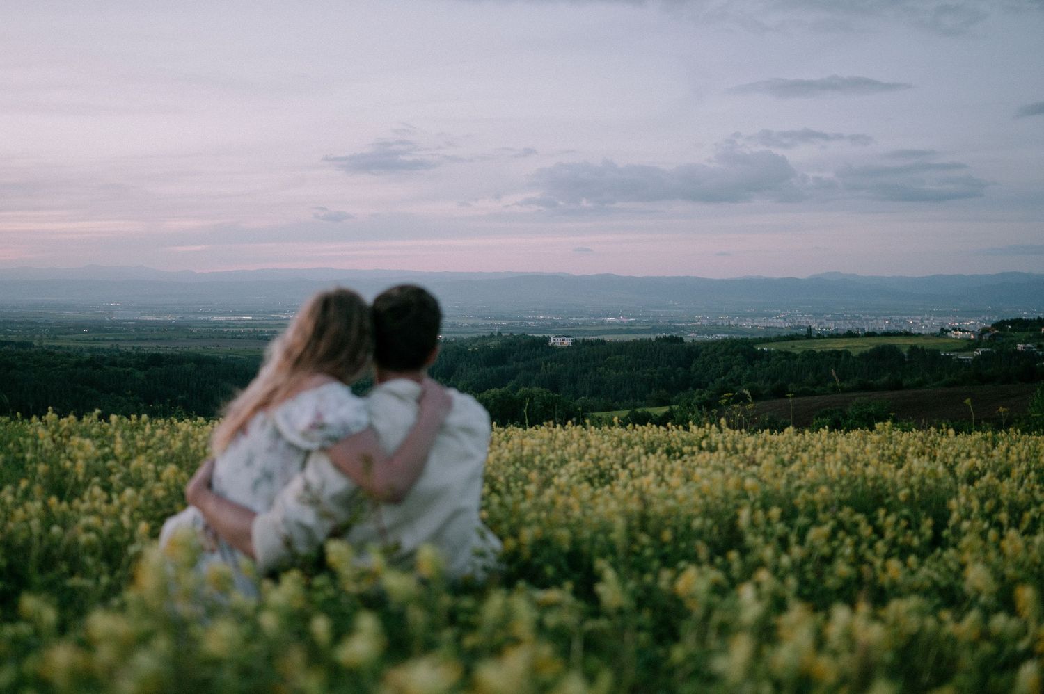 Panoramic view of a couple embracing in a vast yellow flower field at sunset.