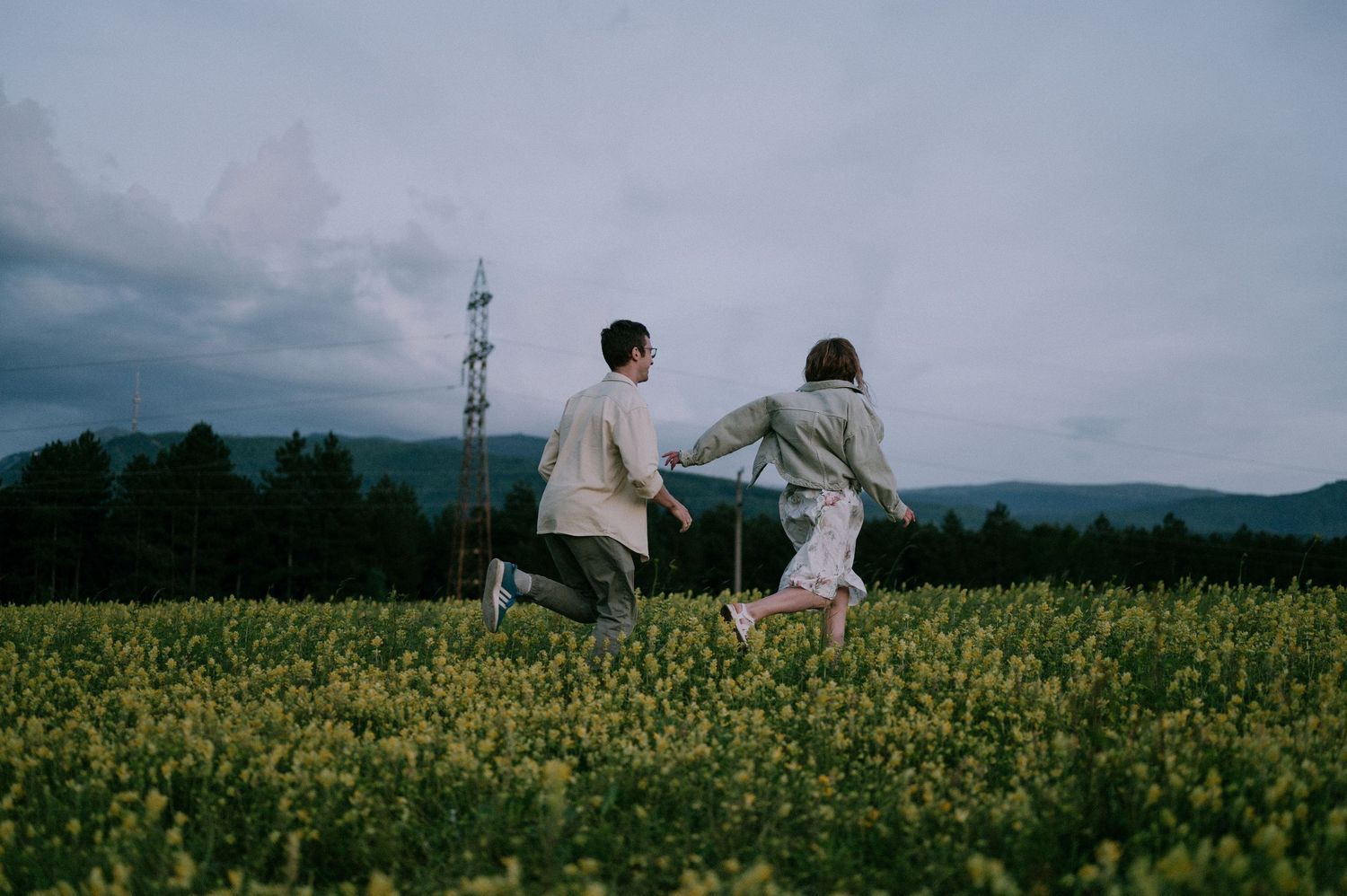 A couple walks through a scenic yellow flower field with mountains in the background.