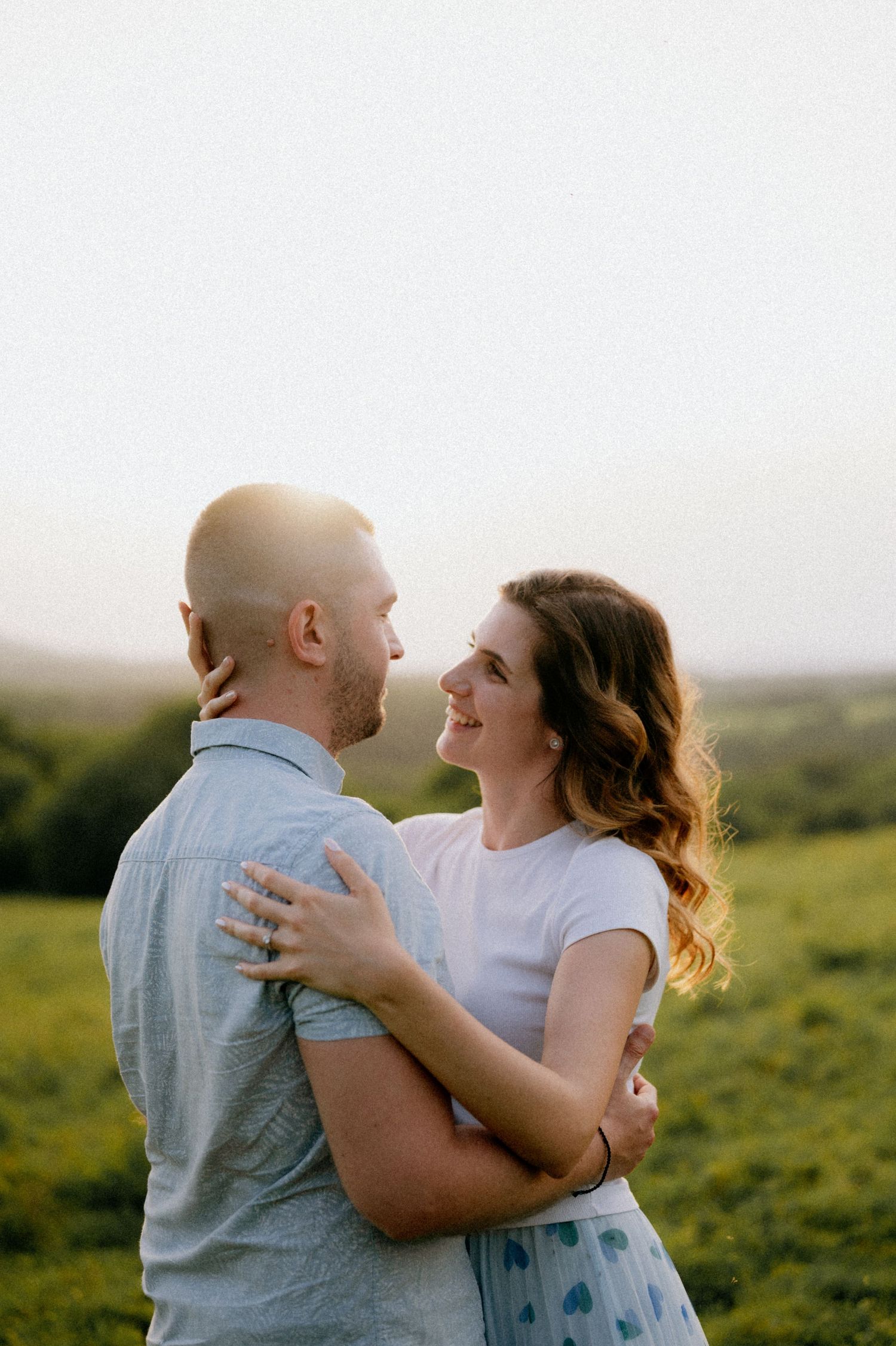 Couple sharing a tender moment in a sunlit meadow with rolling hills in background.
