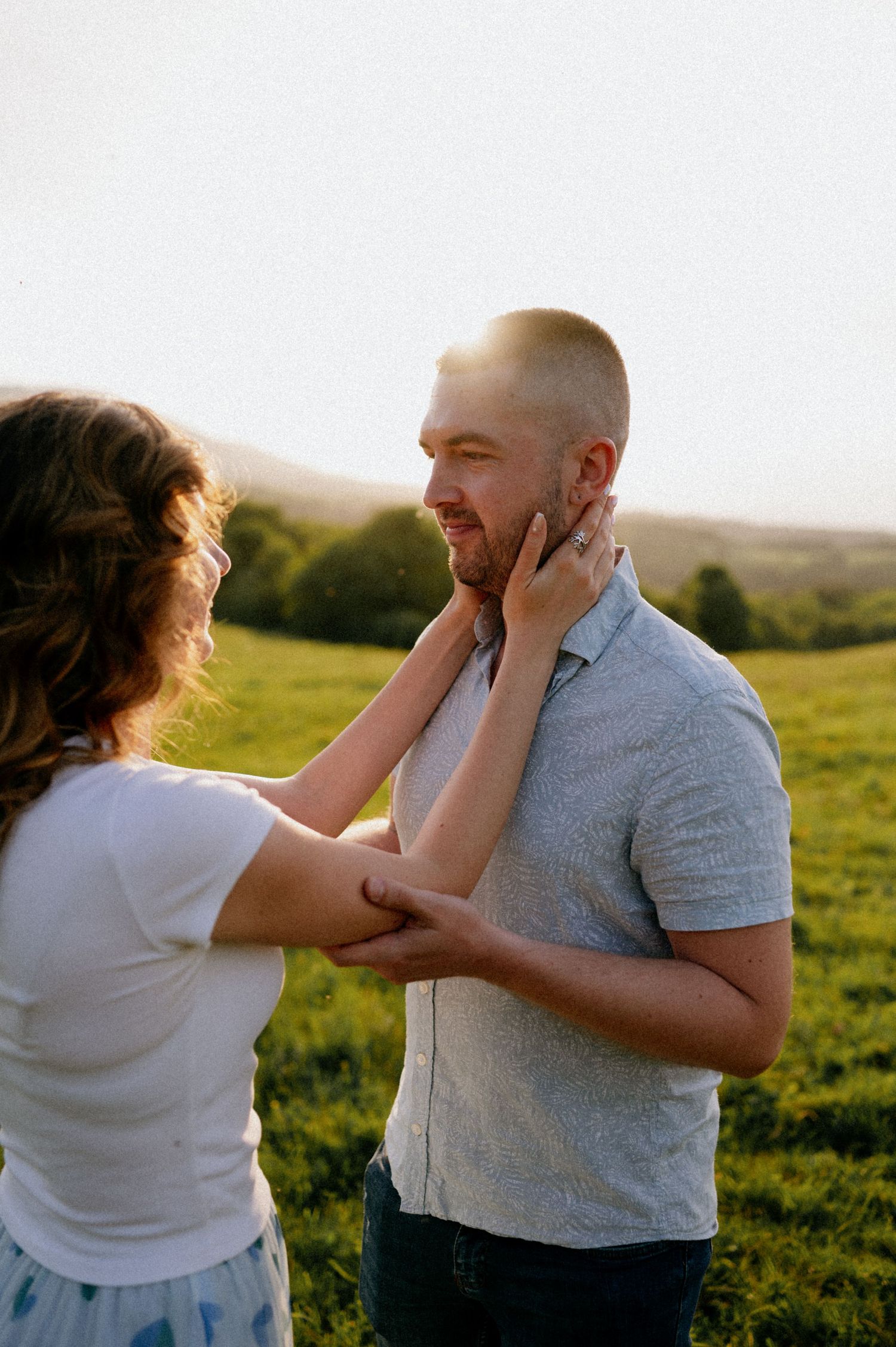 A playful couple shares an intimate moment in a sunlit meadow while holding each other's faces.
