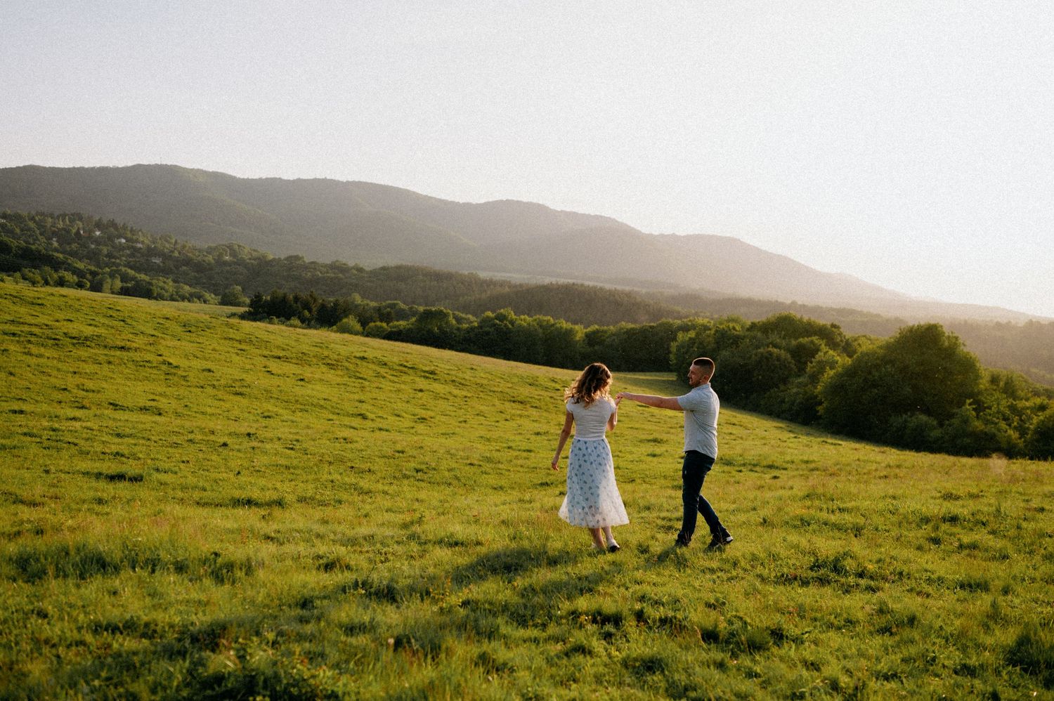 Two people walk hand in hand across a rolling green hillside at dusk.