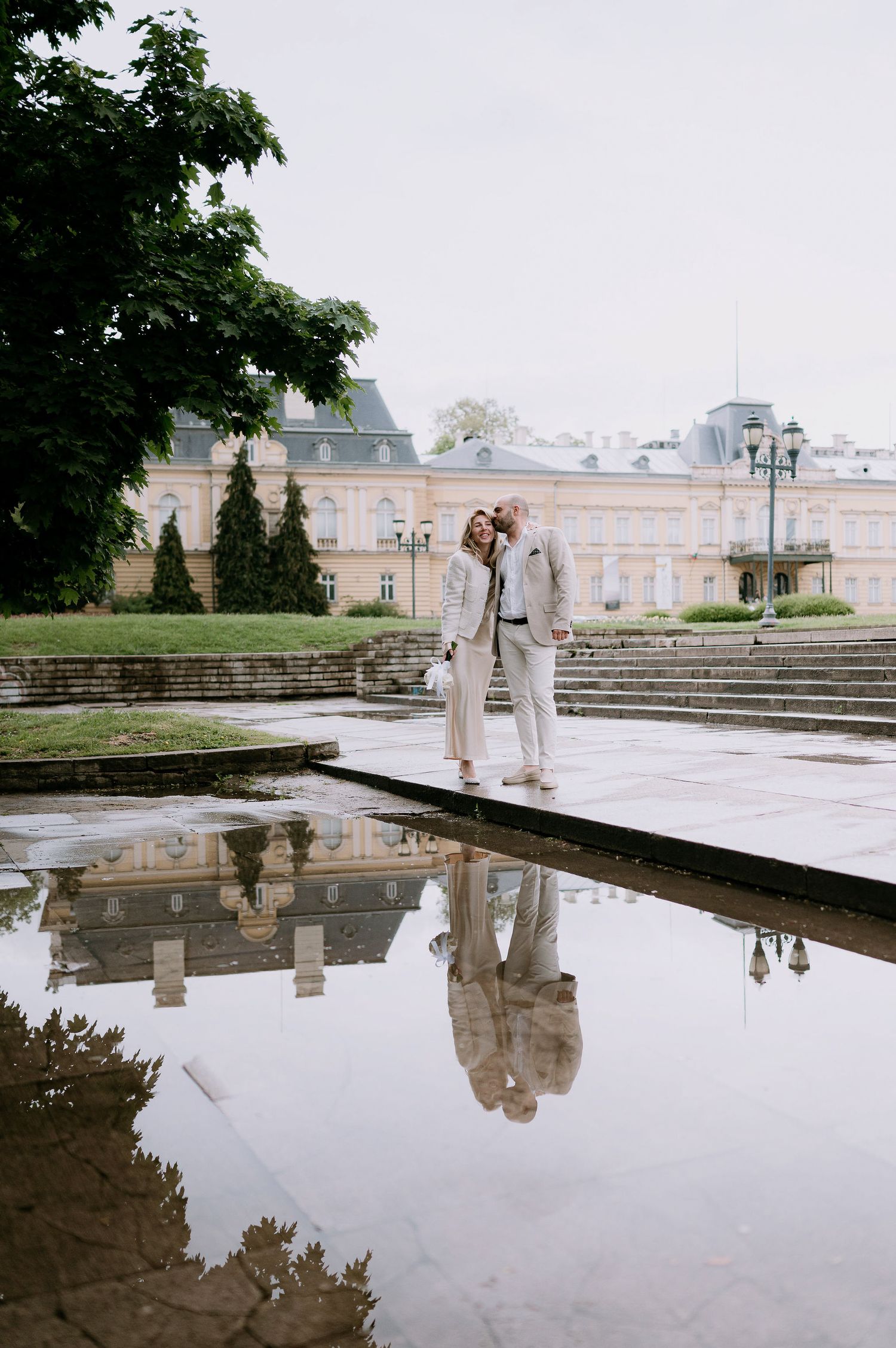 Romantic dance scene reflected in water with elegant architecture and stone steps in background.