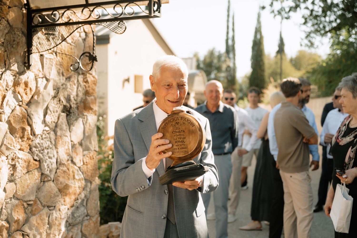 A person in a light gray suit holds a woven basket at an outdoor gathering with cypress trees in the background.