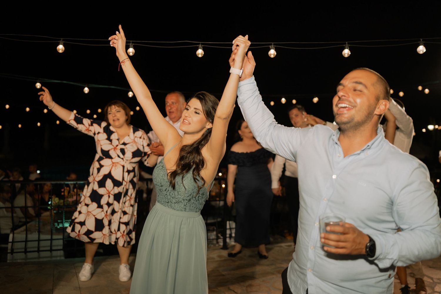 Wedding guests dance joyfully with raised arms under string lights at an outdoor evening reception celebration.