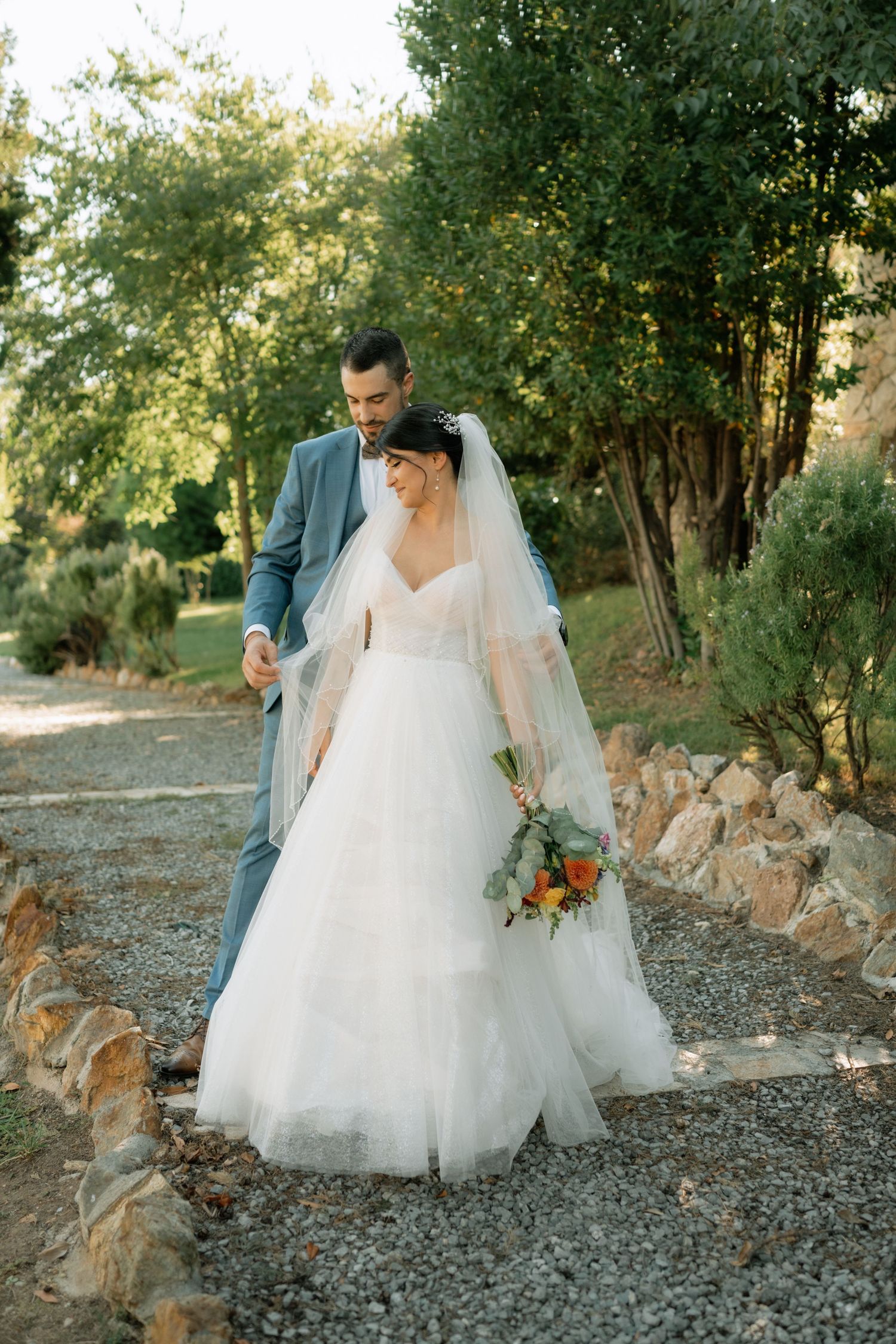 A couple in wedding attire walking together on a gravel path lined with stone walls and trees in golden sunlight.