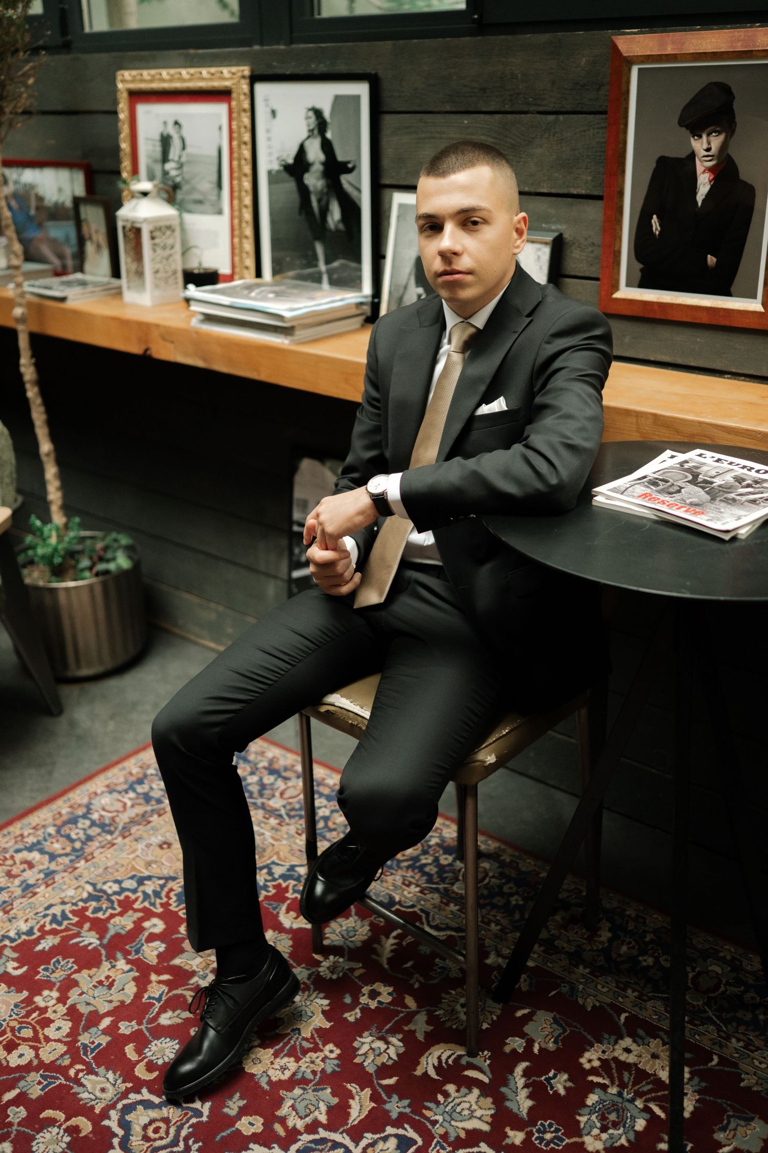 A person in a dark suit sits on a patterned oriental rug in what appears to be an office or formal setting.