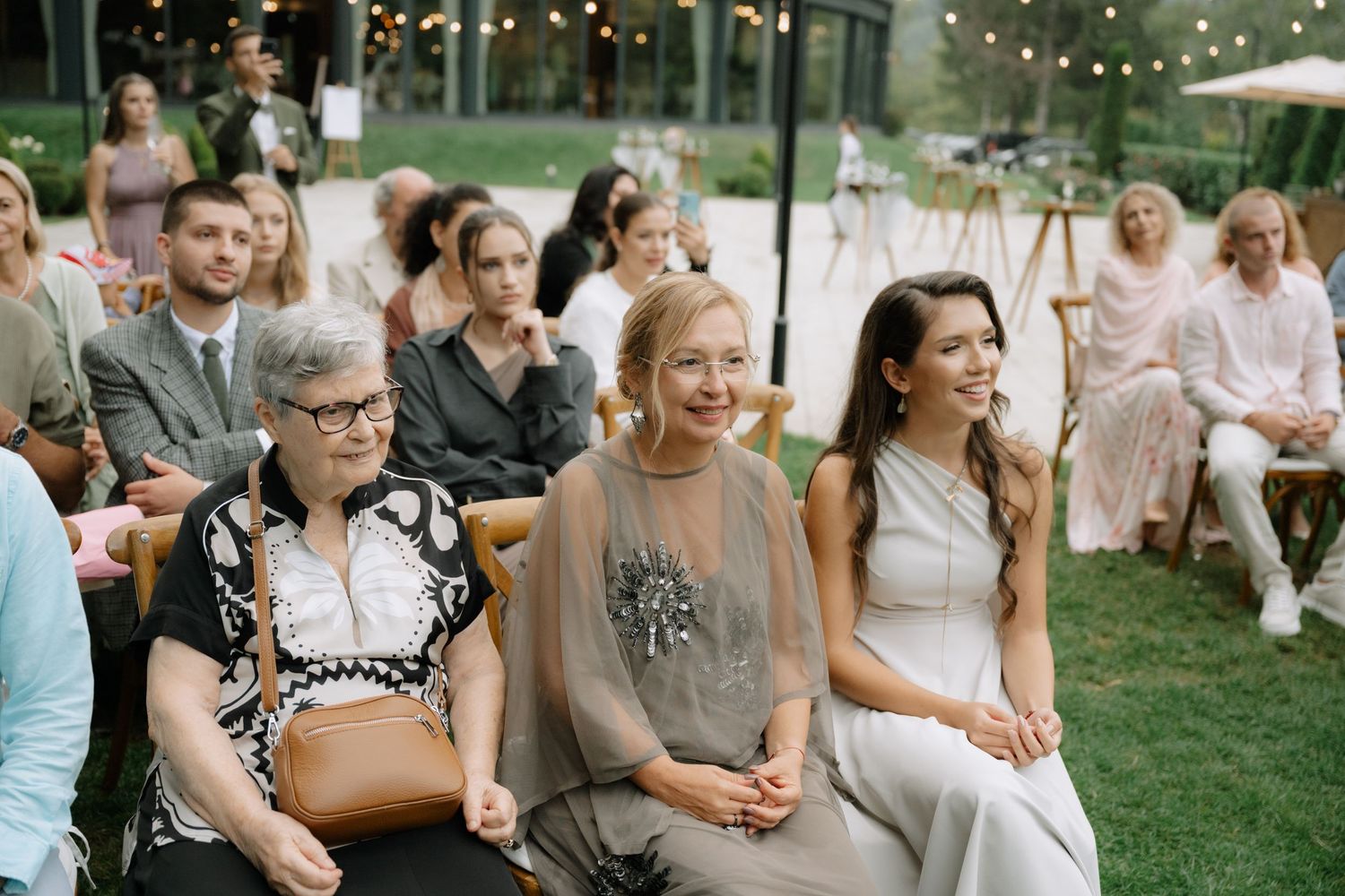 Wedding guests sit on chairs outdoors on a lawn during an evening ceremony with twinkling lights in the background.