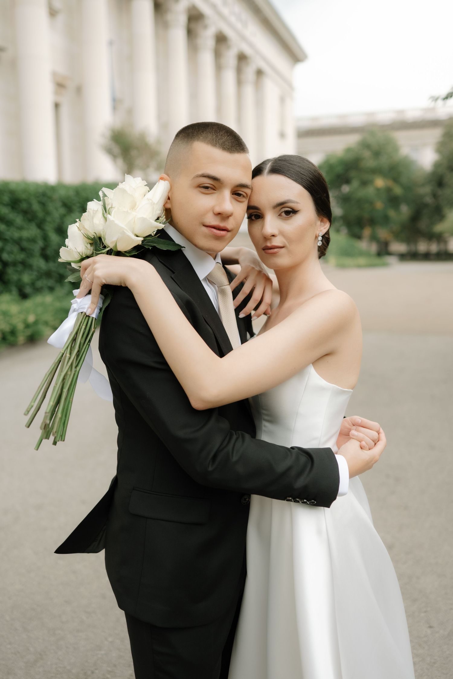 Newlyweds embrace while holding white roses against a classical architectural backdrop.