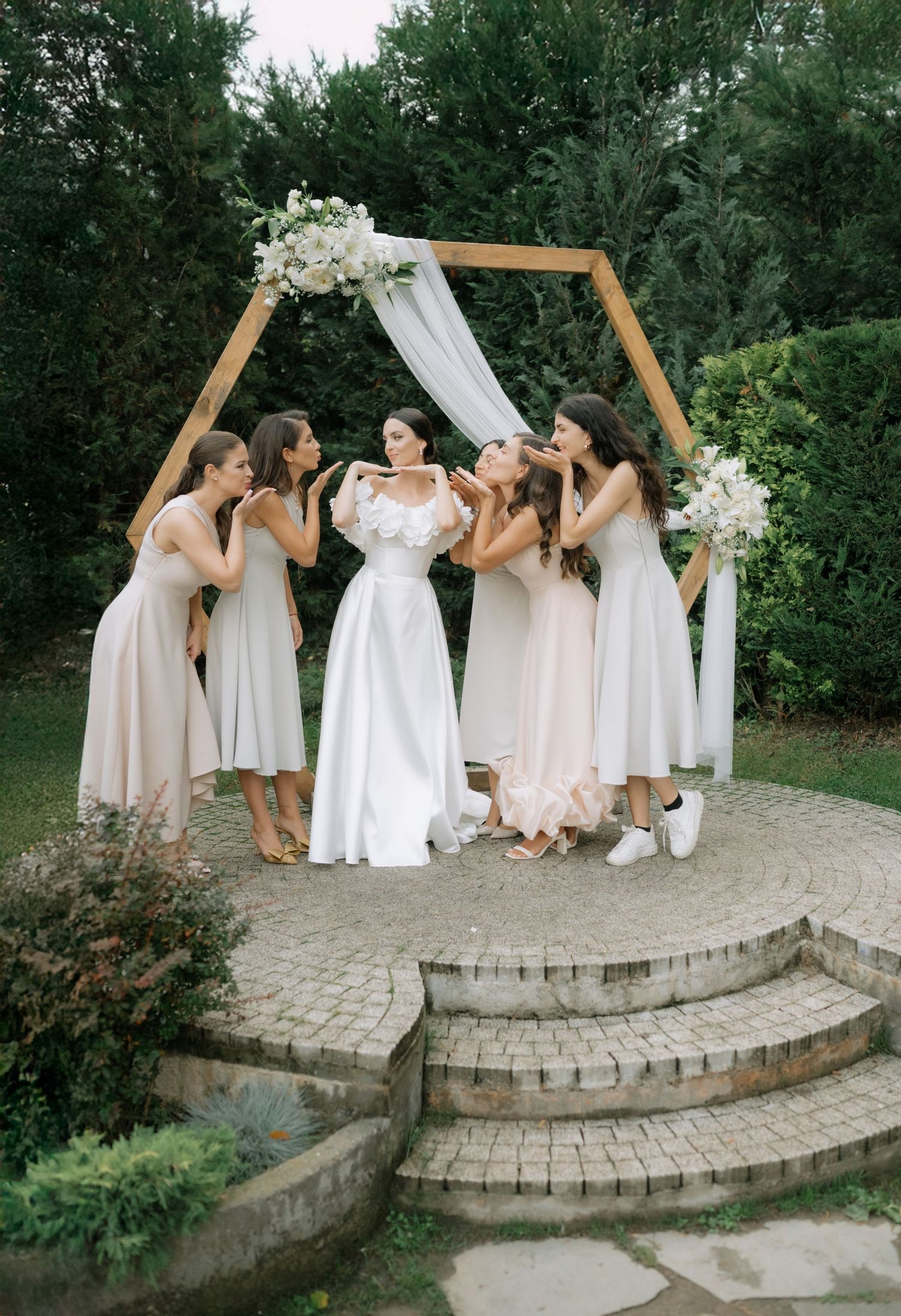 A group in light dresses poses together on curved stone steps surrounded by lush greenery during a wedding celebration.