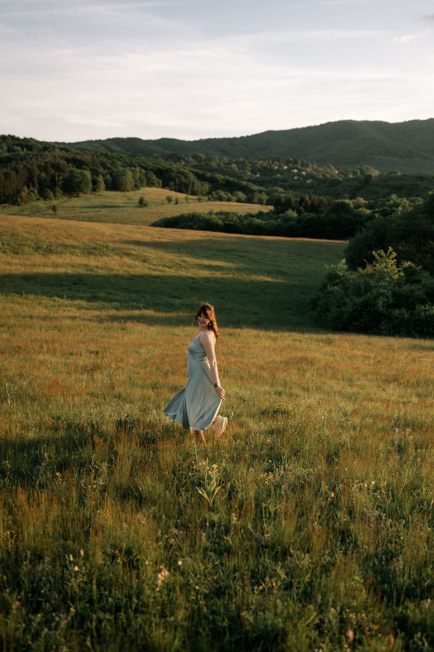 Woman in light blue dress standing in a sunlit green meadow with rolling hills and trees in the background.