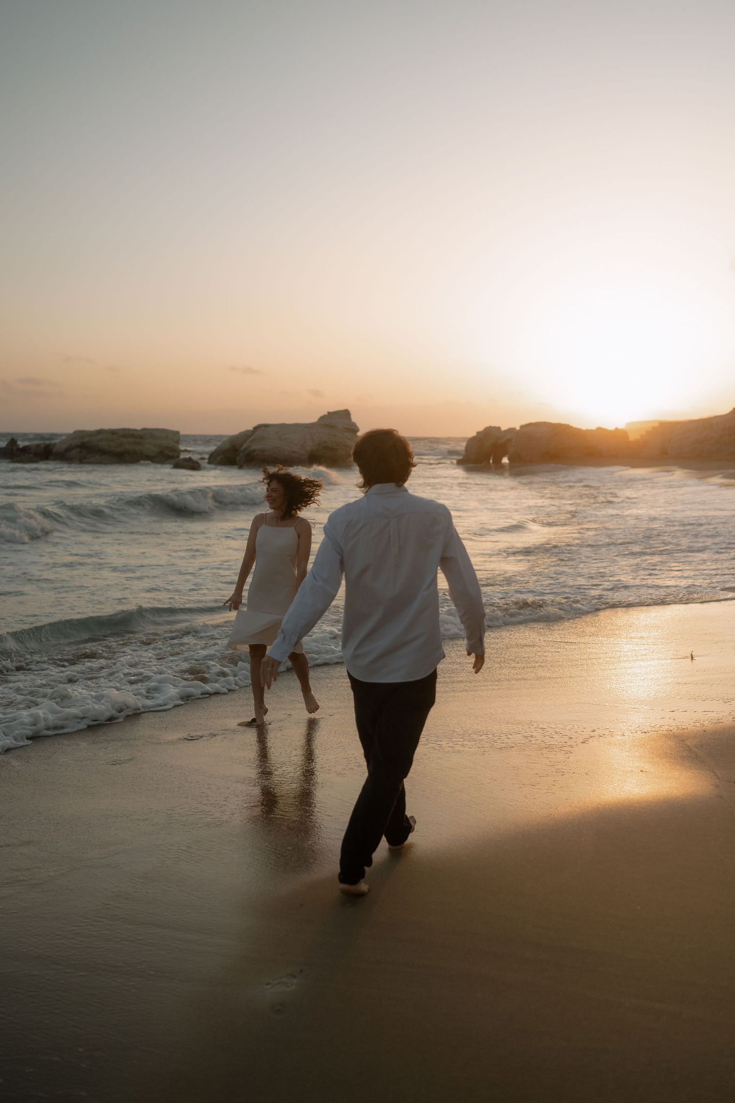 Couple walking hand in hand on a sandy beach at sunset, waves crashing nearby with golden light reflecting on the shore.