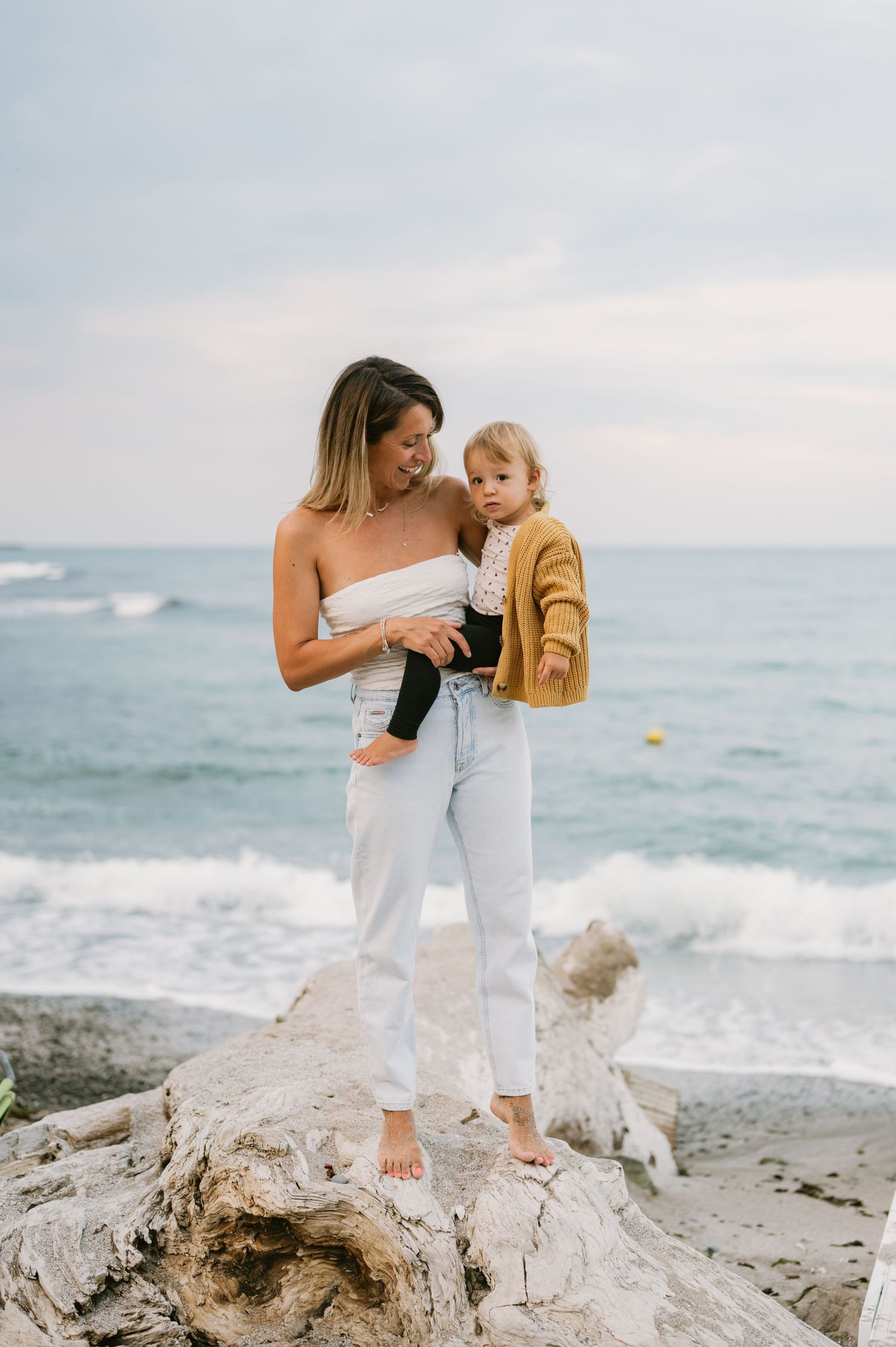 A mother holds her toddler on a rocky beach, waves in the background, wearing white pants and a strapless top.
