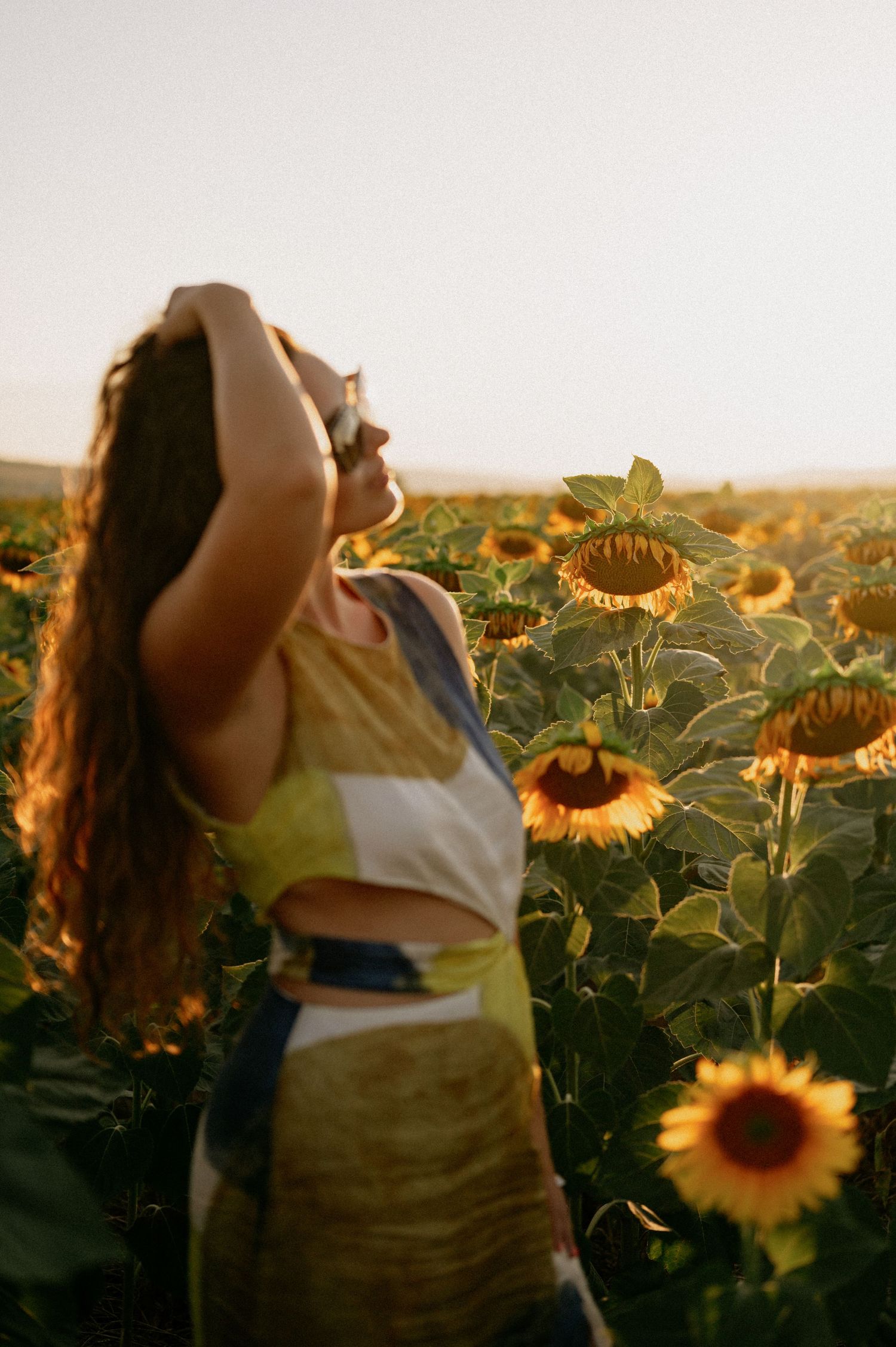 Woman standing in a sunflower field at golden hour, hand raised to her hair, wearing a colorful crop top.