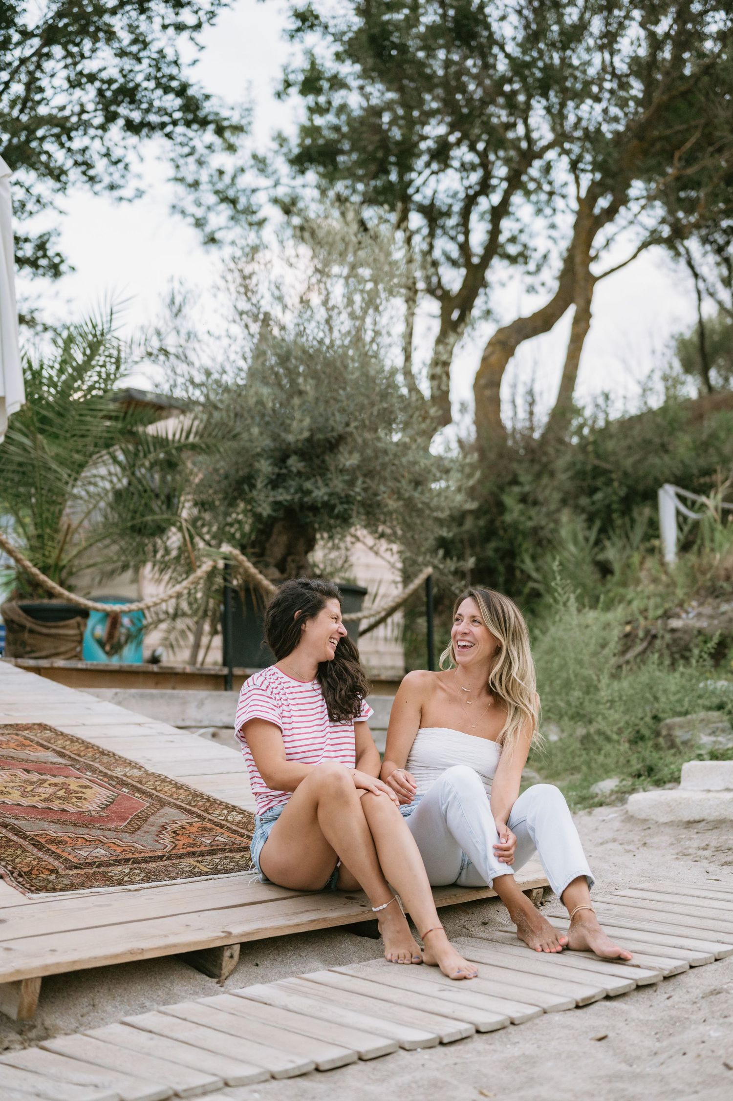 Two women laughing and sitting together on a wooden deck surrounded by trees and a colorful rug in a garden setting.