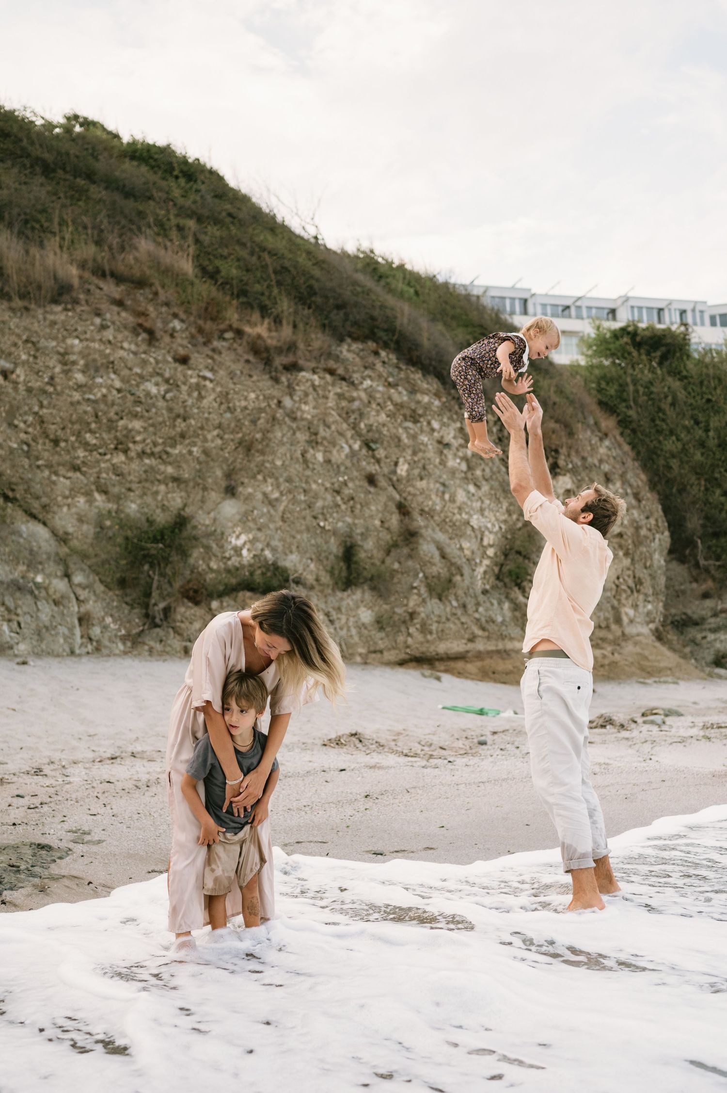Family playing on a sandy beach, father tossing baby in the air while mother hugs young child near rocky cliffs.