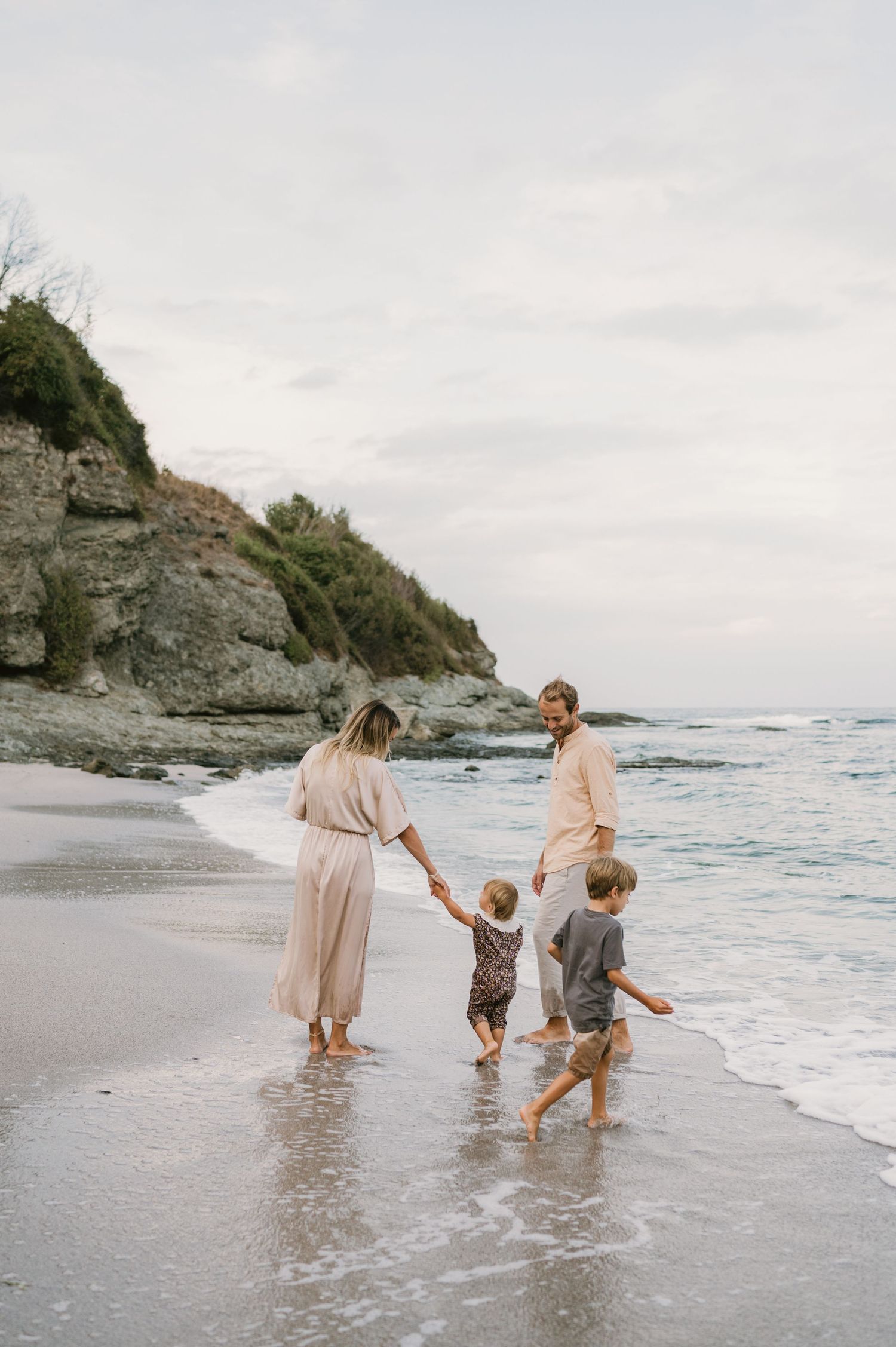 Family of four walking along a sandy beach shoreline, holding hands near rocky cliffs and ocean waves.