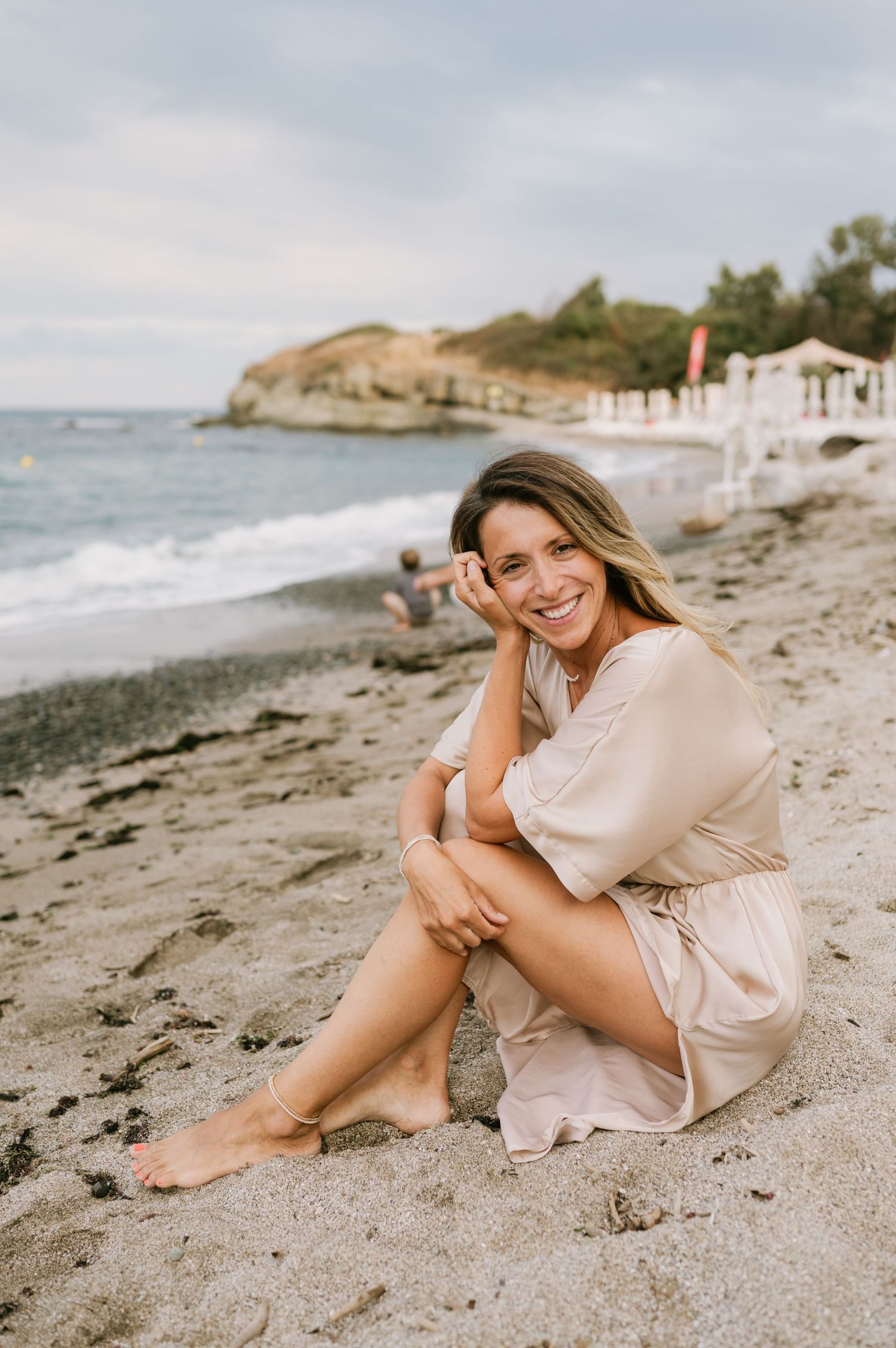 Smiling woman in beige dress sitting barefoot on sandy beach with ocean waves and rocky coastline in background.