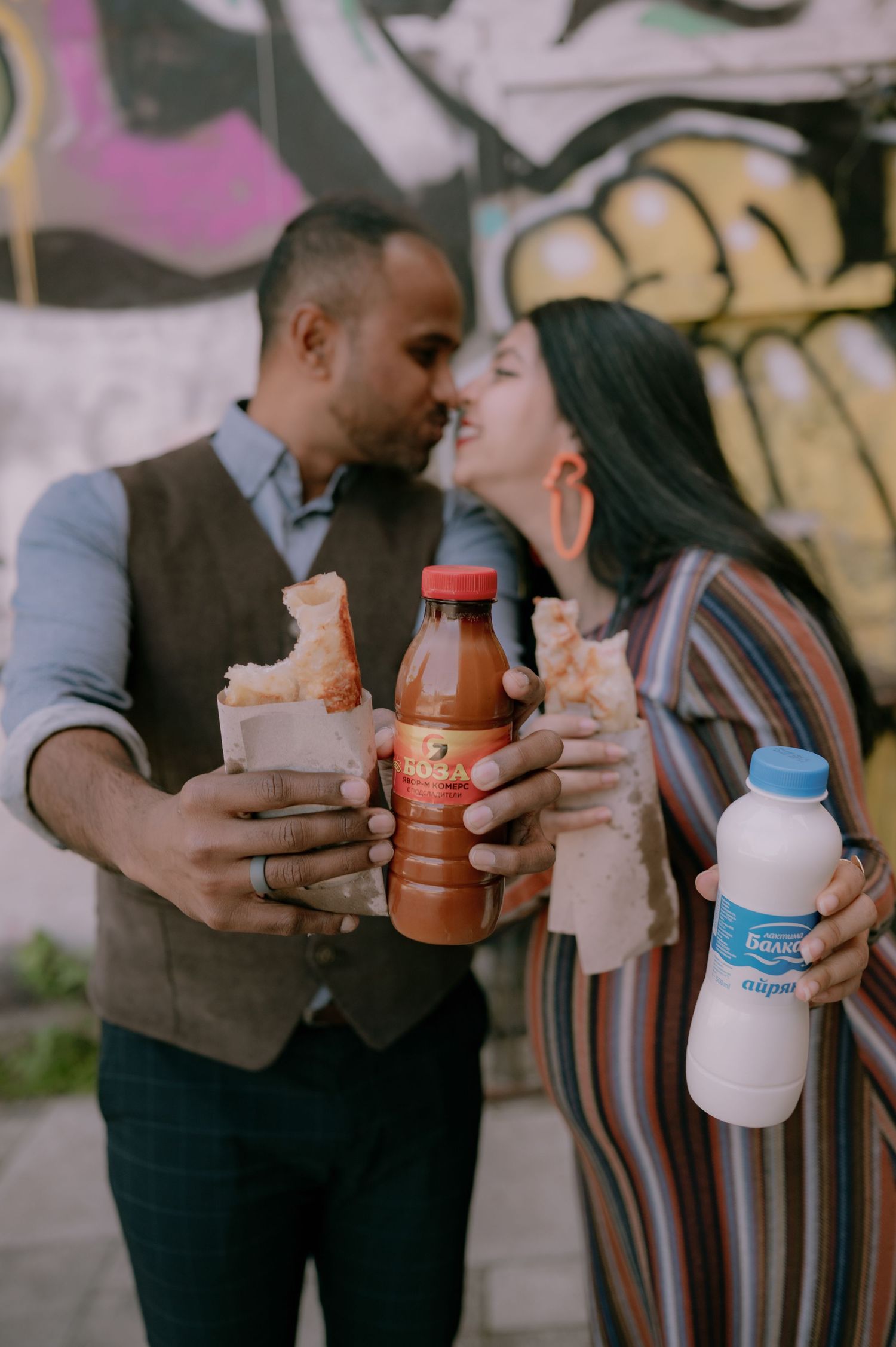 Couple sharing a romantic moment outdoors, holding sandwiches and beverages in front of colorful graffiti wall.
