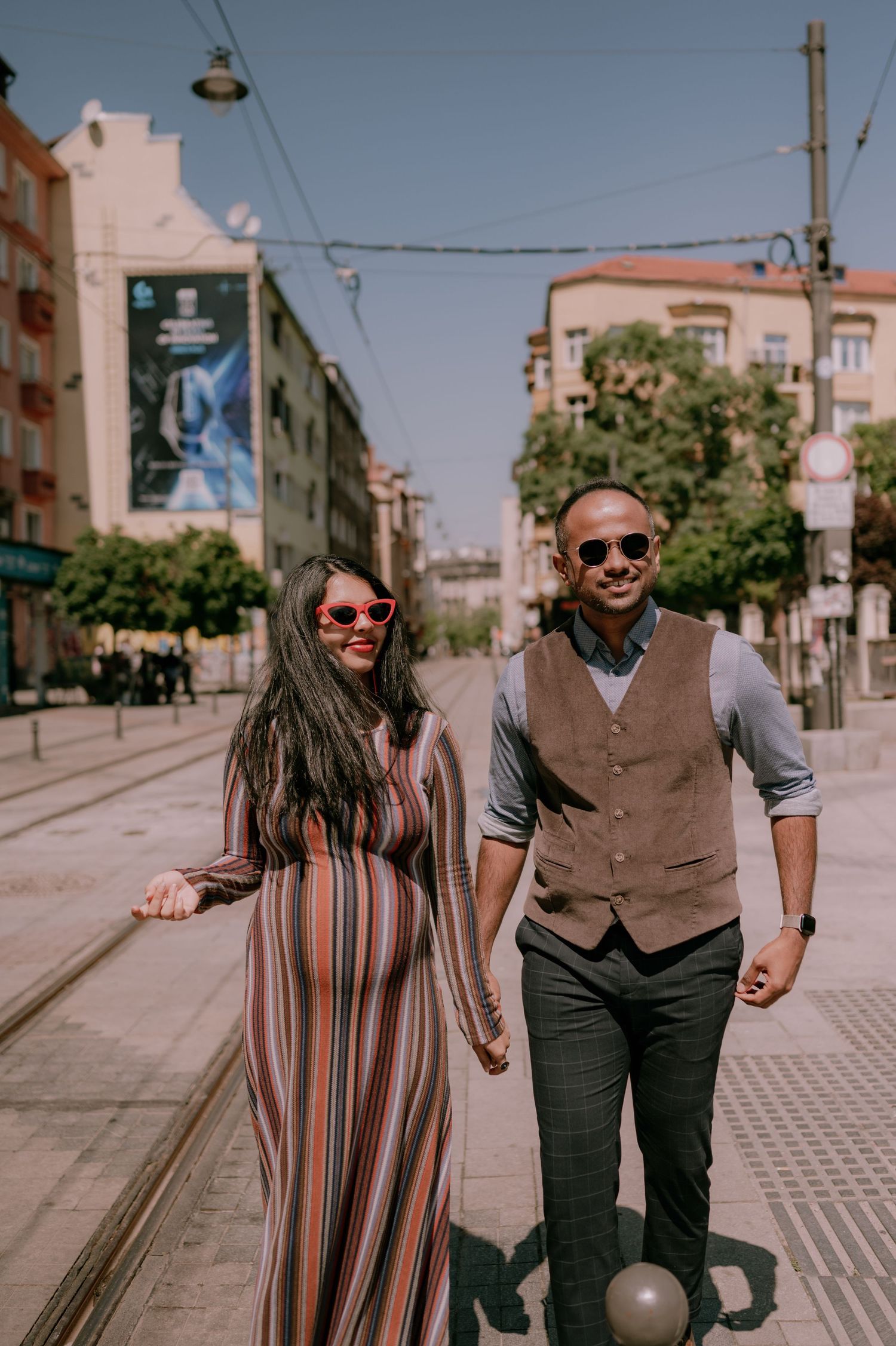 A couple holding hands and walking on a sunny city street, wearing sunglasses and stylish outfits.