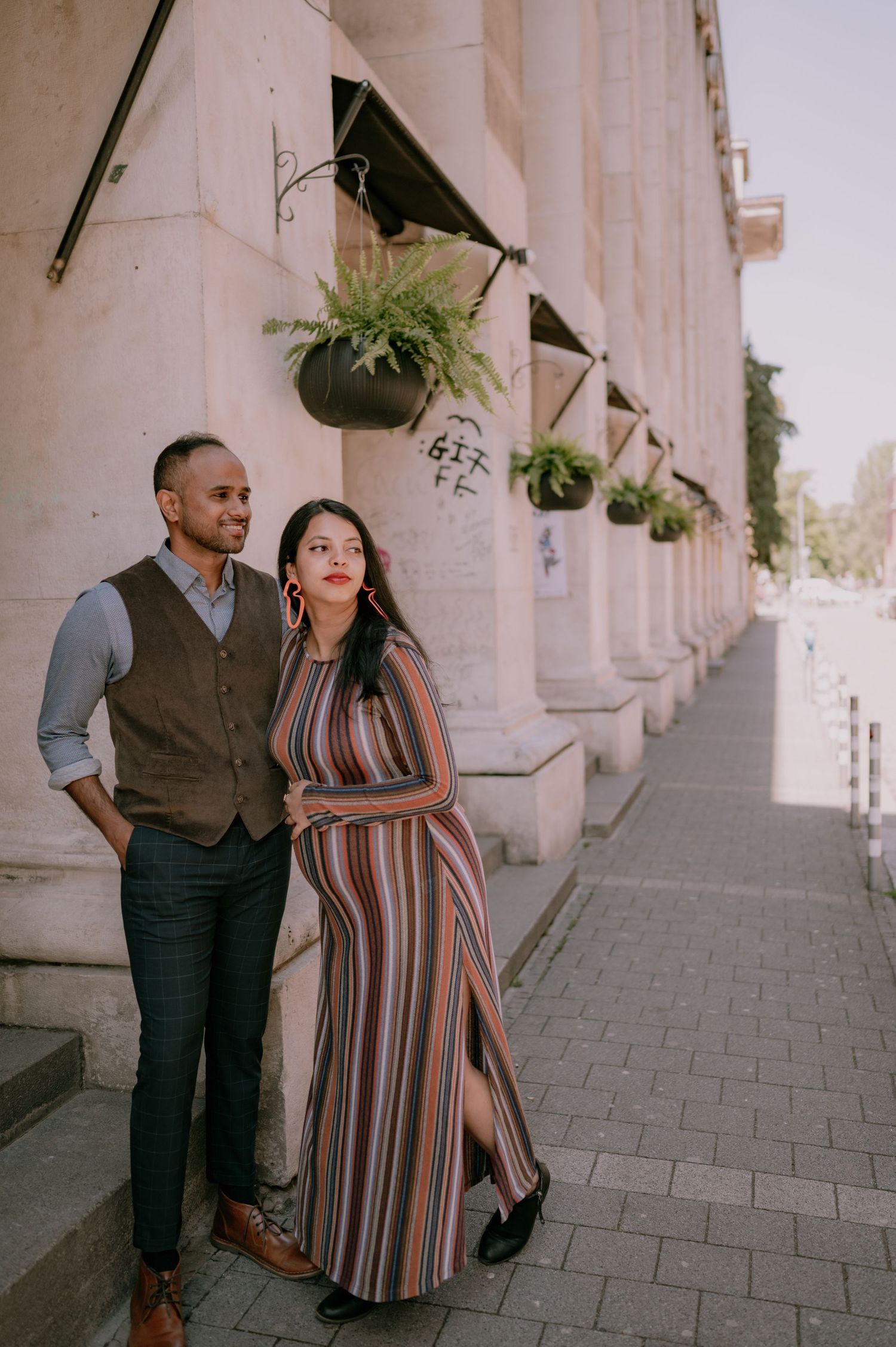 Couple posing on a sidewalk beside a classical building with hanging fern planters, man in vest and woman in striped maxi dress.