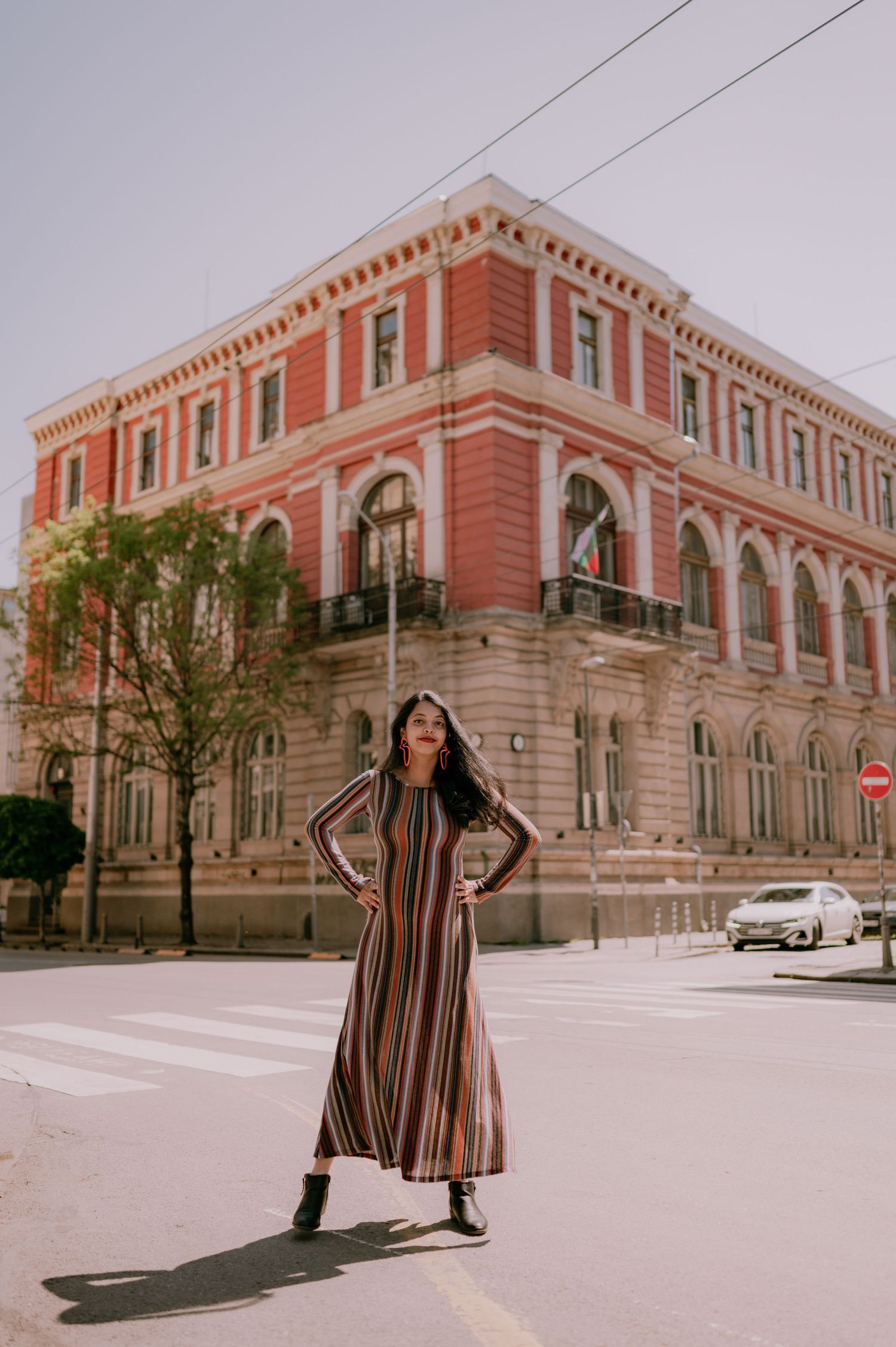 Woman in striped maxi dress posing on street corner before a grand red historic building on a sunny day.