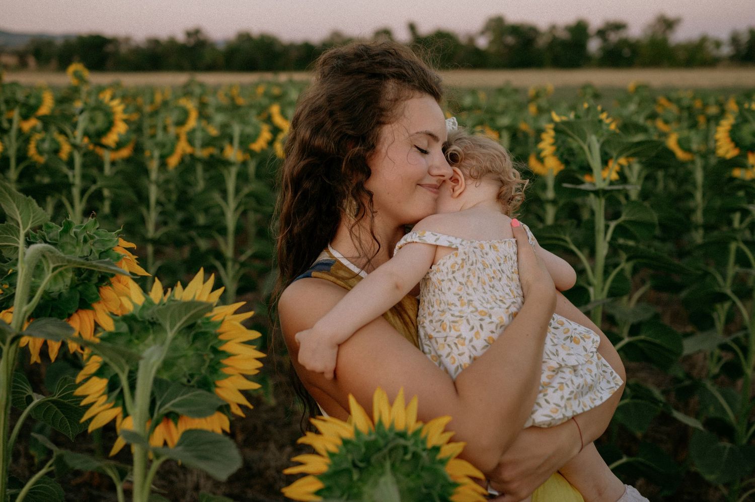 Mother holding baby girl in sunflower field at sunset, sharing a tender moment surrounded by blooming yellow sunflowers.