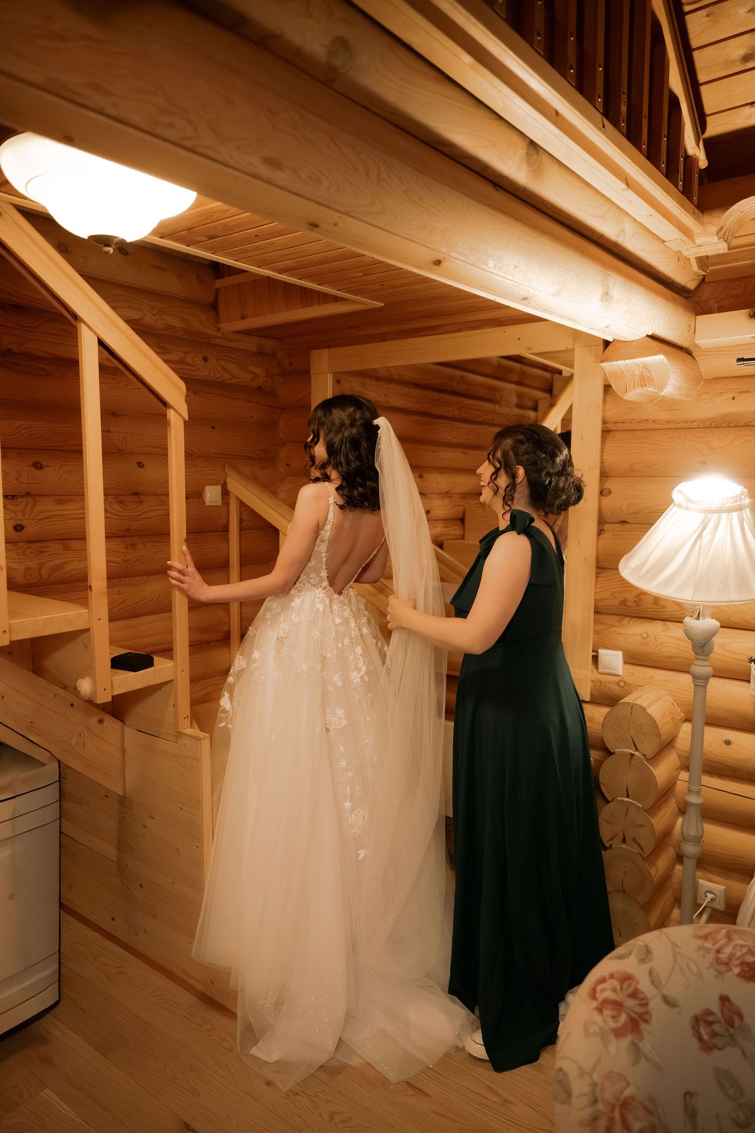 Bride in lace wedding gown with veil assisted by bridesmaid in green dress inside rustic log cabin.