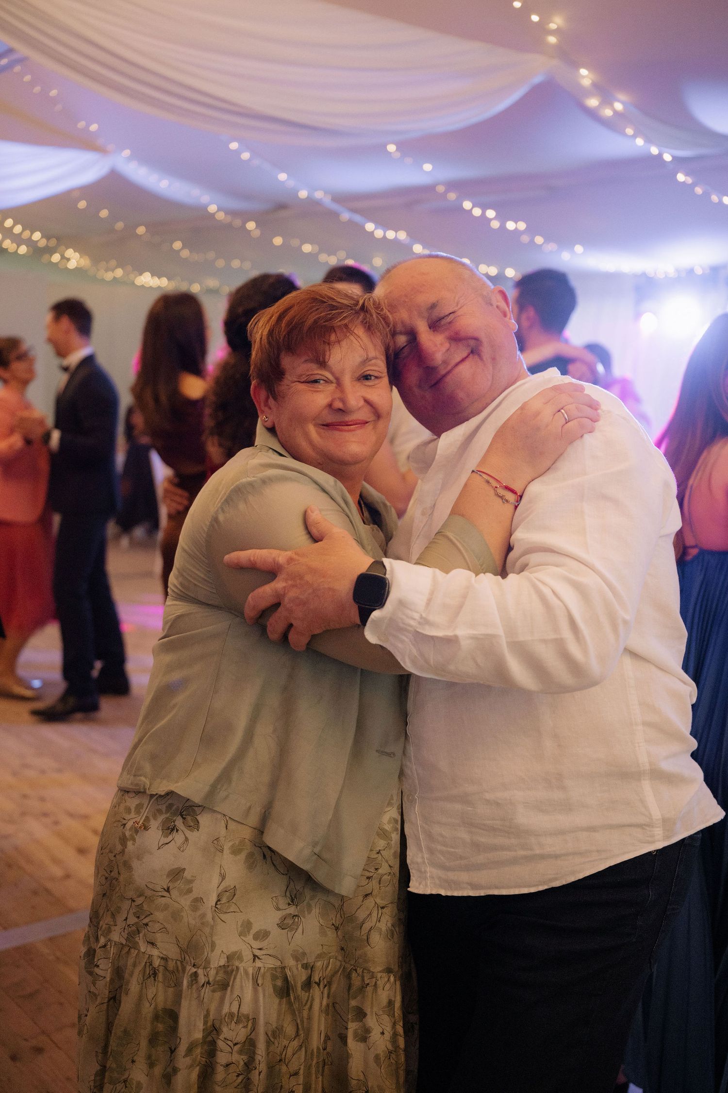 Couple embracing and smiling on a decorated wedding dance floor with fairy lights and other guests dancing in background.