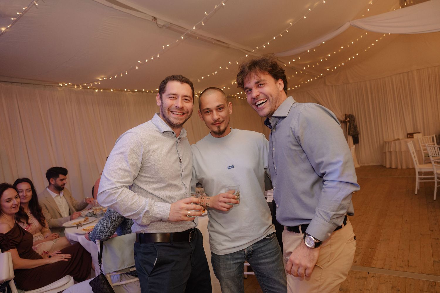 Three smiling men pose together at an elegant indoor event with string lights and white draping.