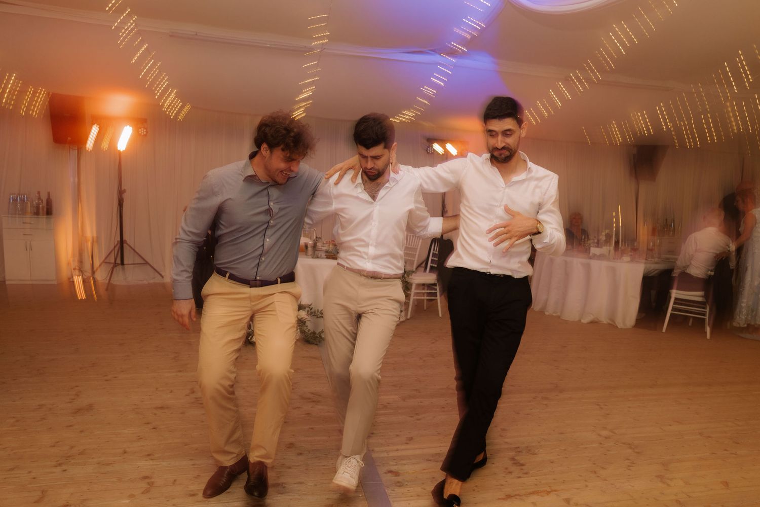 Three men dancing together at a wedding reception in a warmly lit banquet hall.