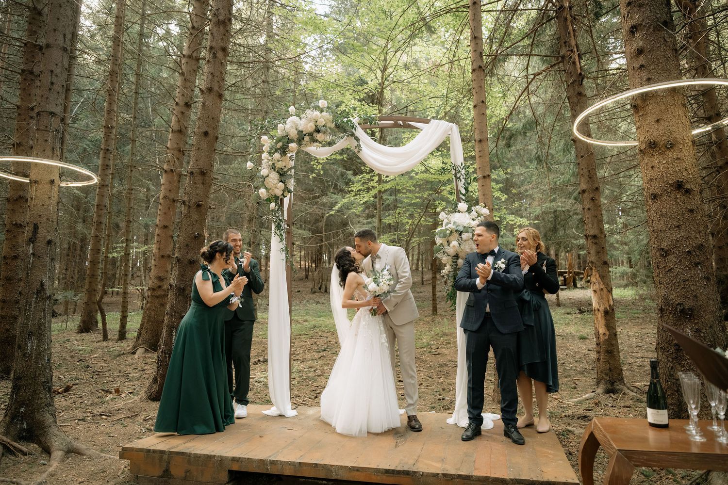 Couple kissing at outdoor forest wedding ceremony with floral arch, bridesmaids in green dresses, and guests celebrating.