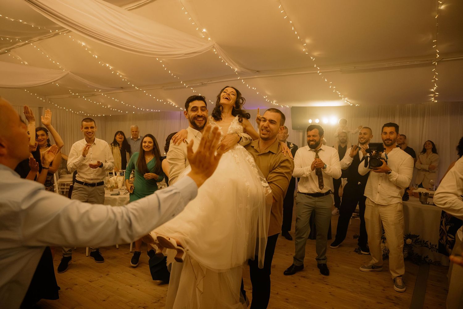 Bride lifted by groom on wedding dance floor surrounded by celebrating guests in elegant fairy light decorated venue.