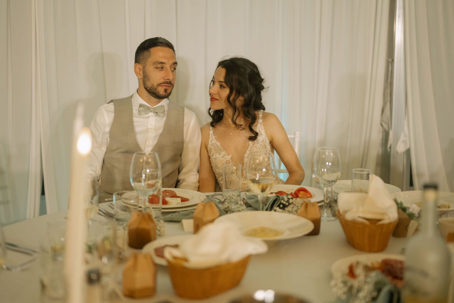 Elegant couple in wedding attire seated at a beautifully decorated reception table with candles and glassware.