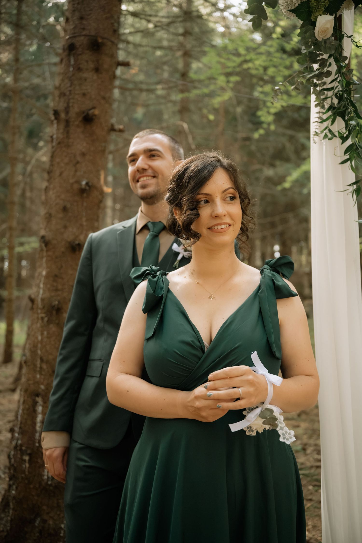 Couple in matching emerald green wedding attire posing in a forest ceremony setting with floral arch.