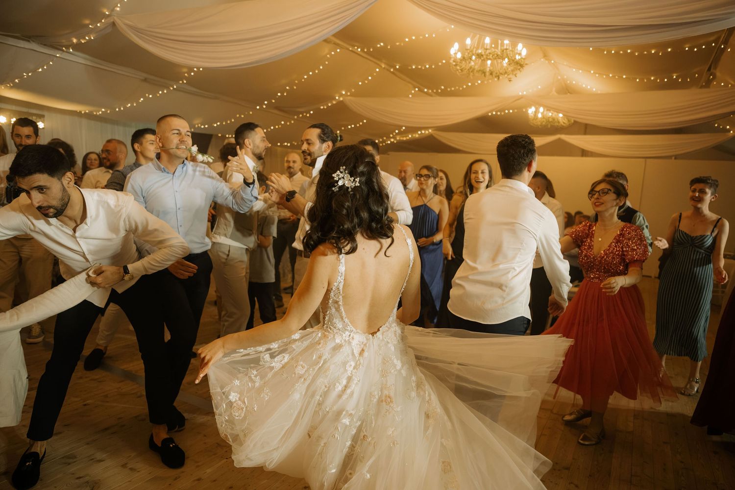 Bride and groom dance joyfully with wedding guests under warm string lights in elegant reception venue.