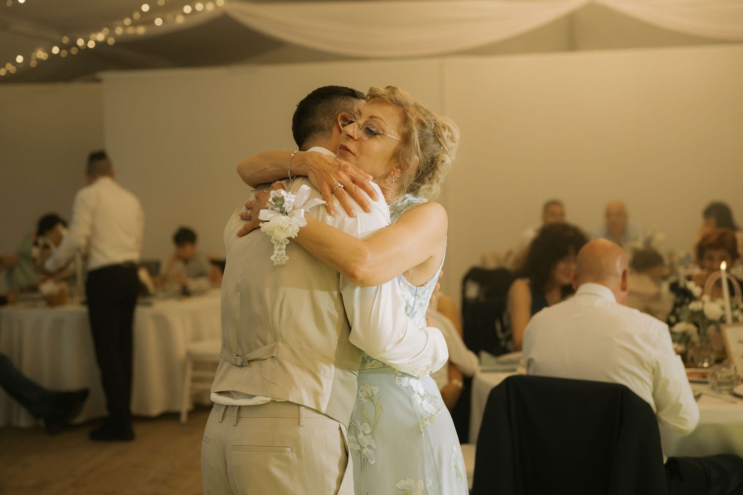 Couple sharing a tender first dance embrace at a wedding reception with guests seated at decorated tables in background.
