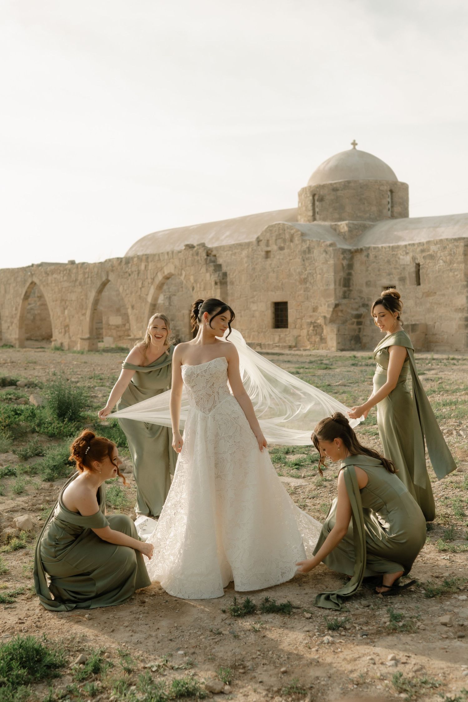 Bride in white gown with bridesmaids in olive green dresses arranging veil near ancient stone ruins.