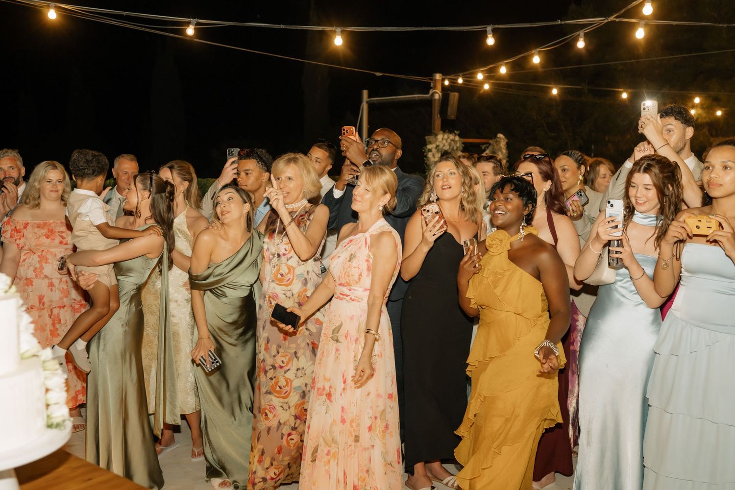 Wedding guests in colorful dresses dancing and celebrating outdoors at night under string lights.