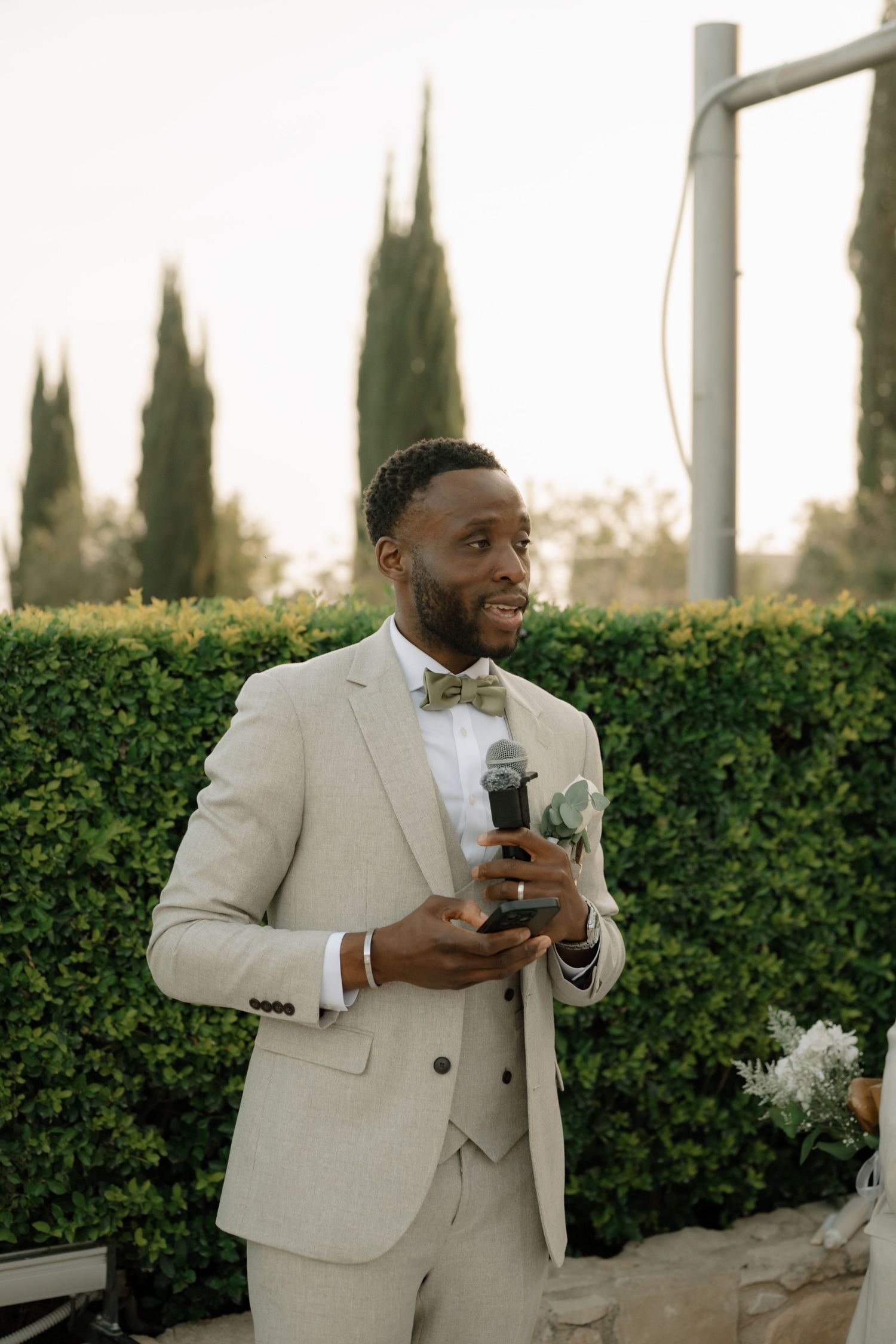 A smiling groom in a beige suit and bow tie holds a microphone while giving a speech at an outdoor wedding ceremony.