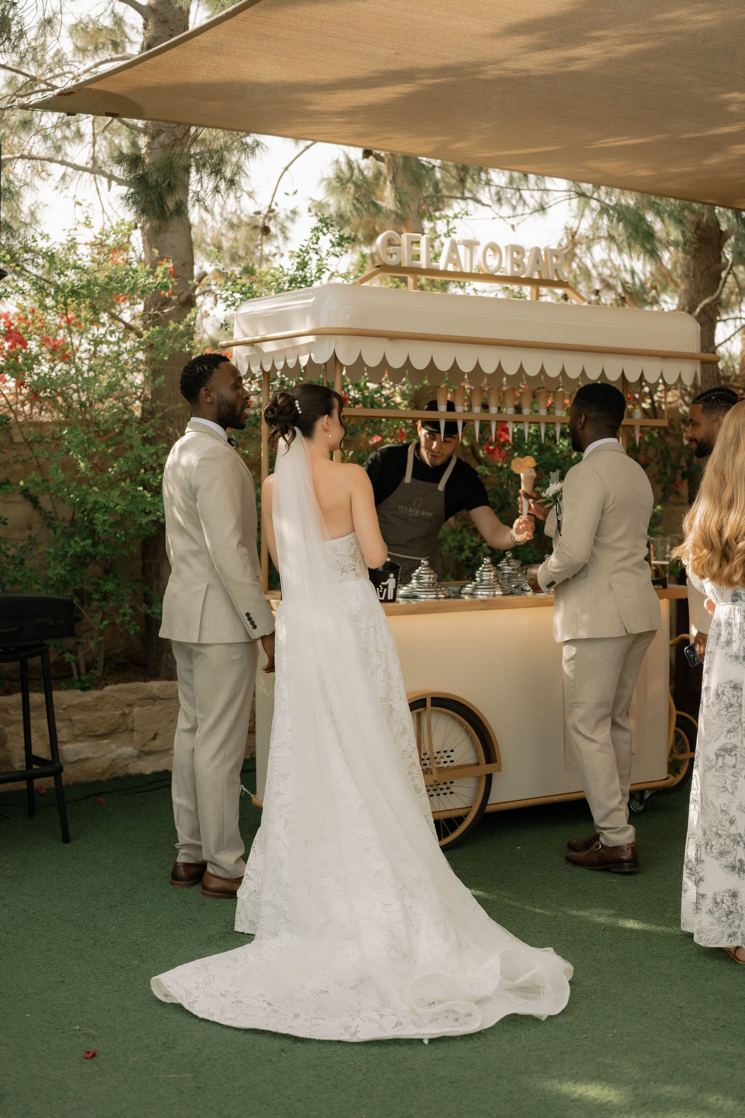 Bride and groom at an outdoor gelato cart during their wedding reception, served by a vendor under a shaded canopy.