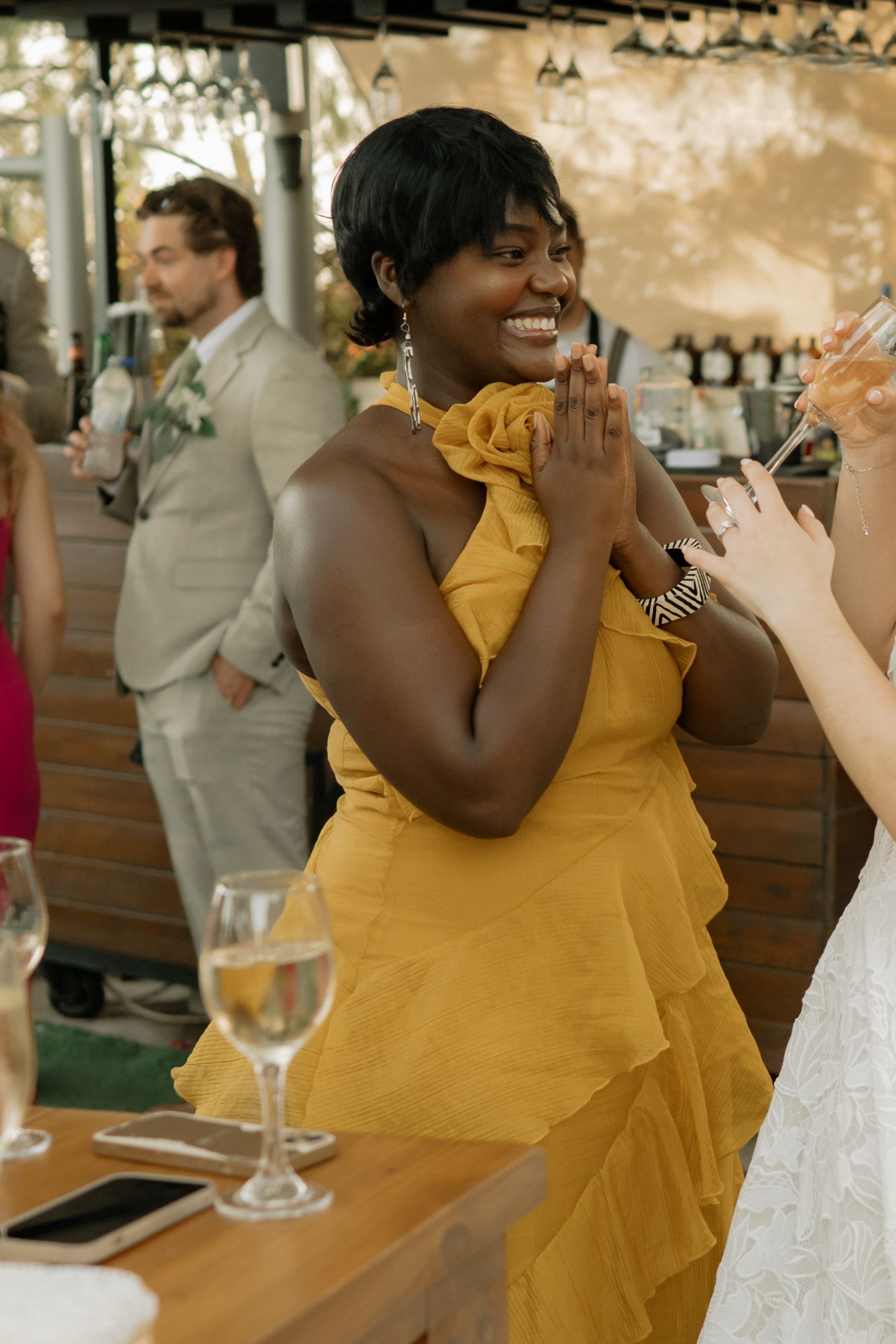 Woman in yellow ruffled dress smiling joyfully at a wedding reception, hands clasped together.