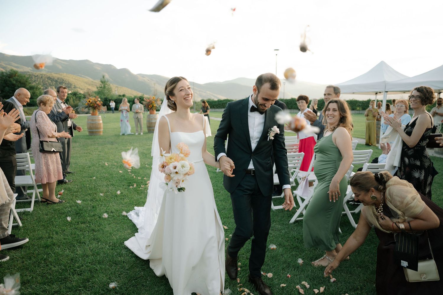 Wedding guests toss flower petals in celebration during an outdoor ceremony in a scenic mountain setting.