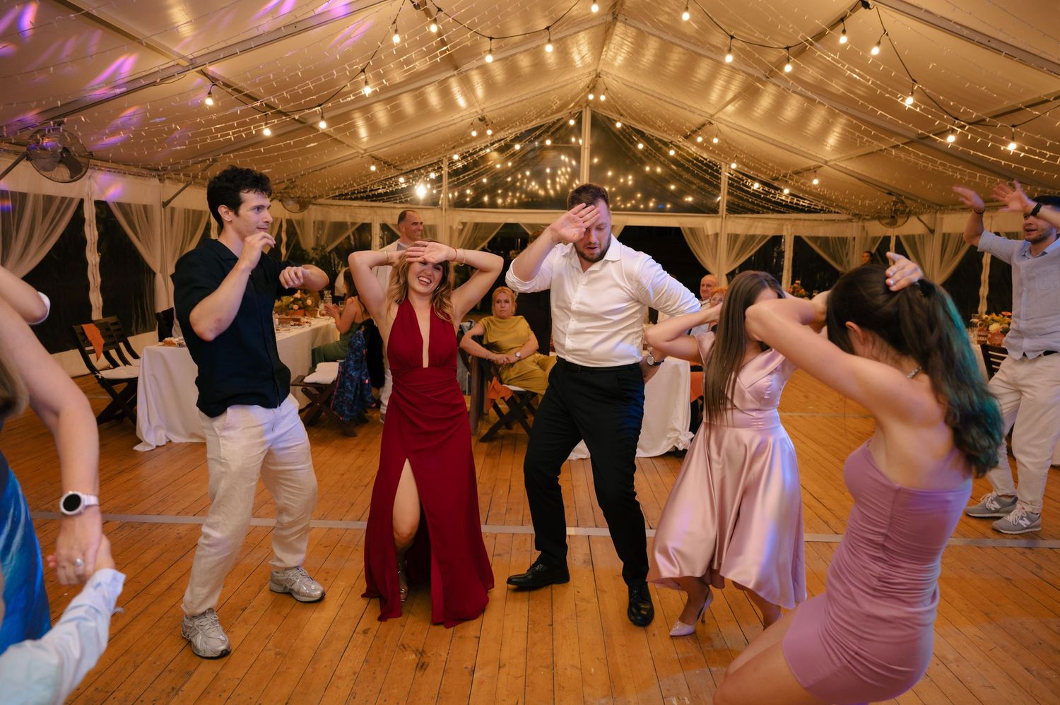 Wedding guests dance enthusiastically on a wooden dance floor under a tent with twinkling lights at an evening reception.