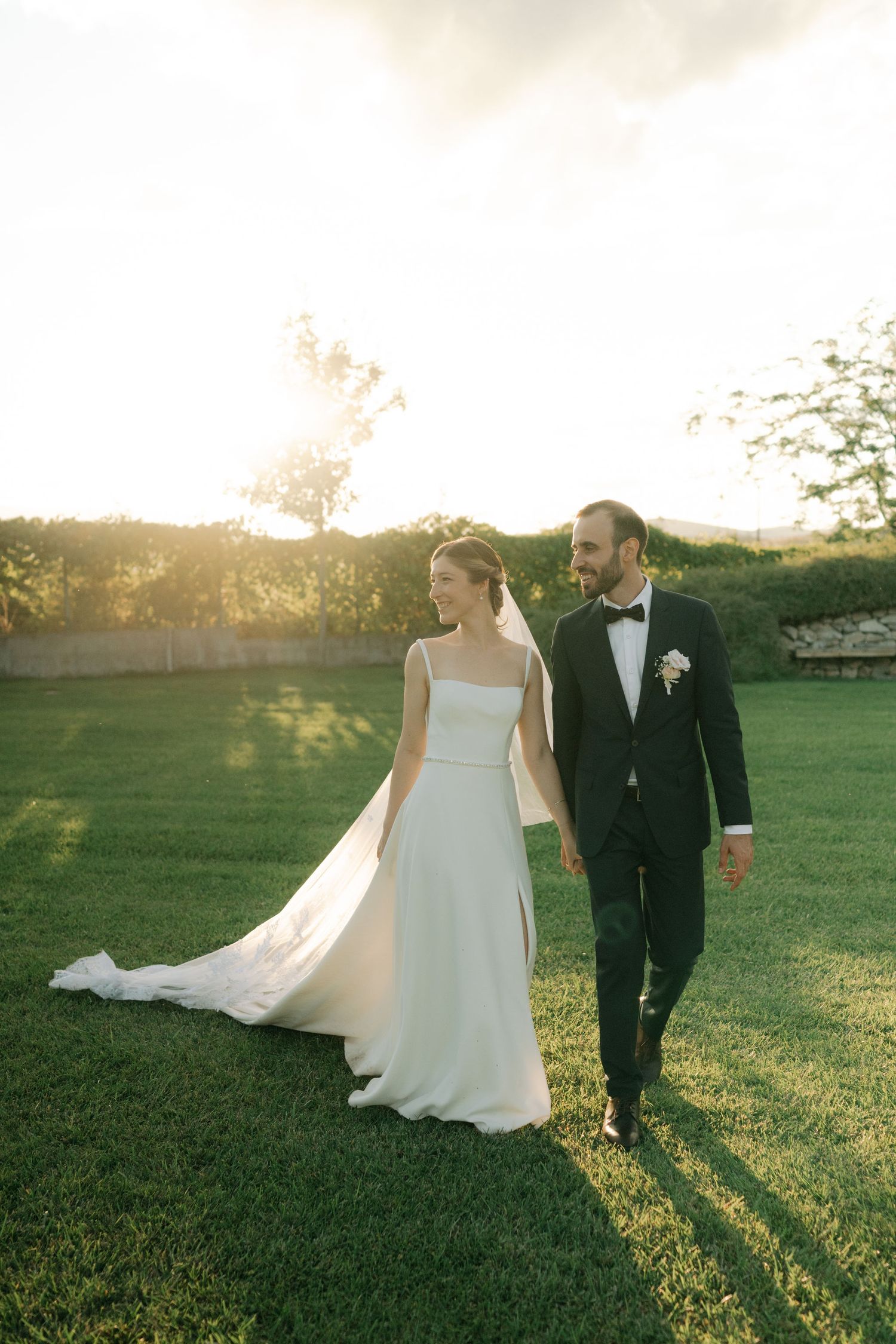 A bride and groom walk hand in hand across a sunlit green field during golden hour at sunset.