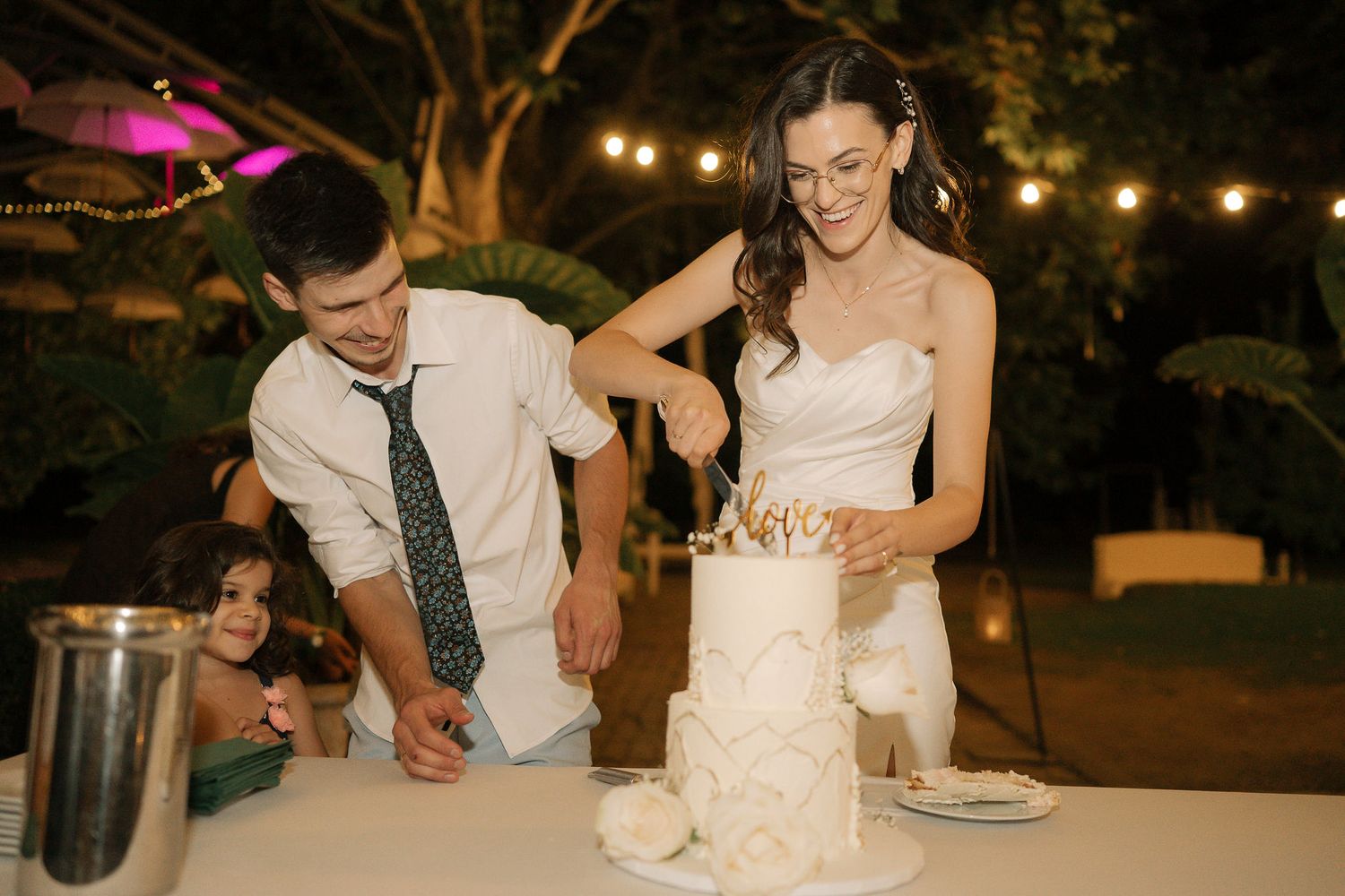 A couple cuts their wedding cake together at an outdoor evening reception with string lights glowing in the background.